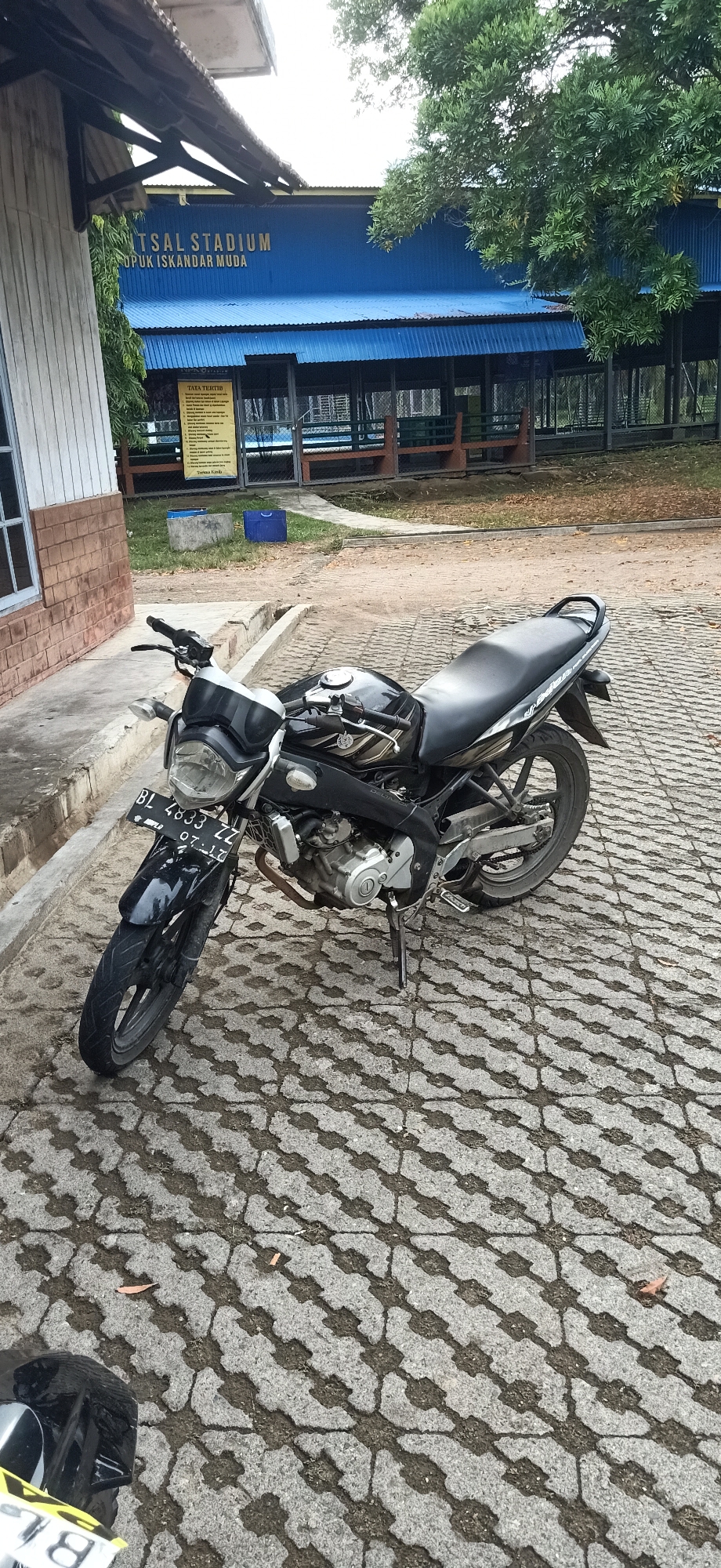 In Palohgadeng, Indonesia, a black motorcycle is parked on an outdoor paved area in front of a building with a blue roof that reads "FUTSAL STADIUM". The building appears to be a sports facility. The motorcycle has a license plate that reads "BL 4833 ZZ" and the date "07-17". There is also a partial view of another motorcycle with a yellow license plate in the bottom left corner of the image. The paved area is made of interlocking gray bricks arranged in a geometric pattern. In the background, there are trees and some fencing around a sports field. The overall scene depicts a sunny day in an outdoor setting.
