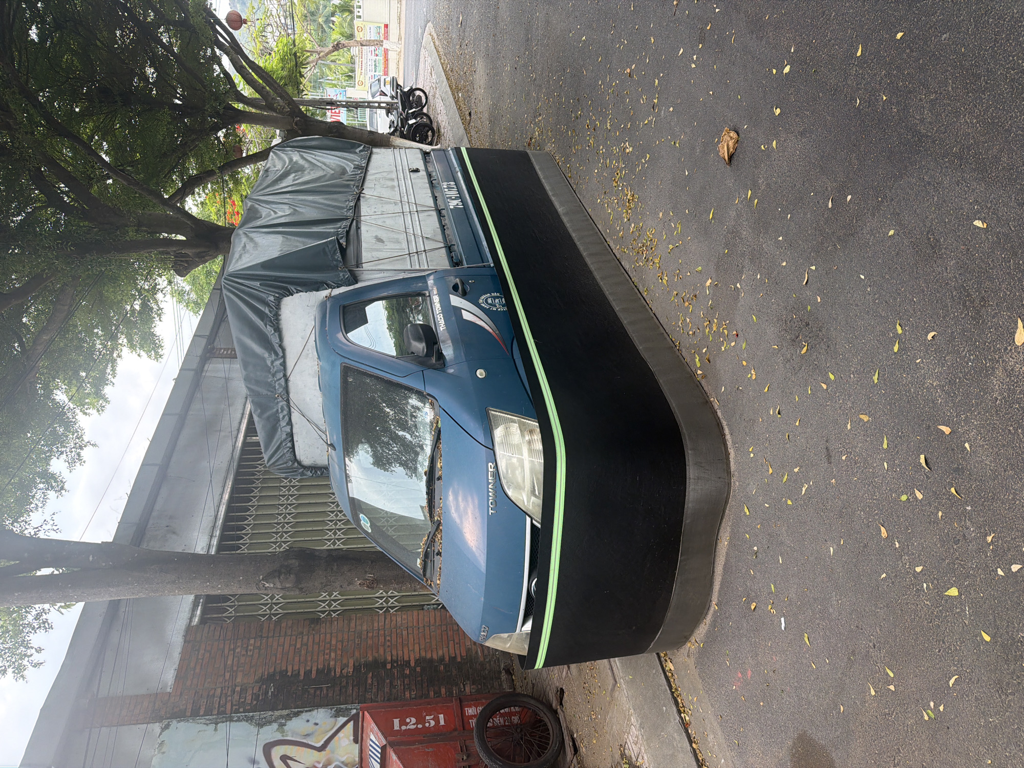 A blue and silver cargo truck with a black lower skirt is parked on a street in Bac Nha Trang Ward, Vietnam. The truck has a tarp covering its cargo bed. The street is asphalt and is covered with fallen leaves. To the left of the truck is a red metal object with the number "12.51" printed on it, and a bicycle wheel. Behind the truck are trees and buildings. The sky is cloudy.