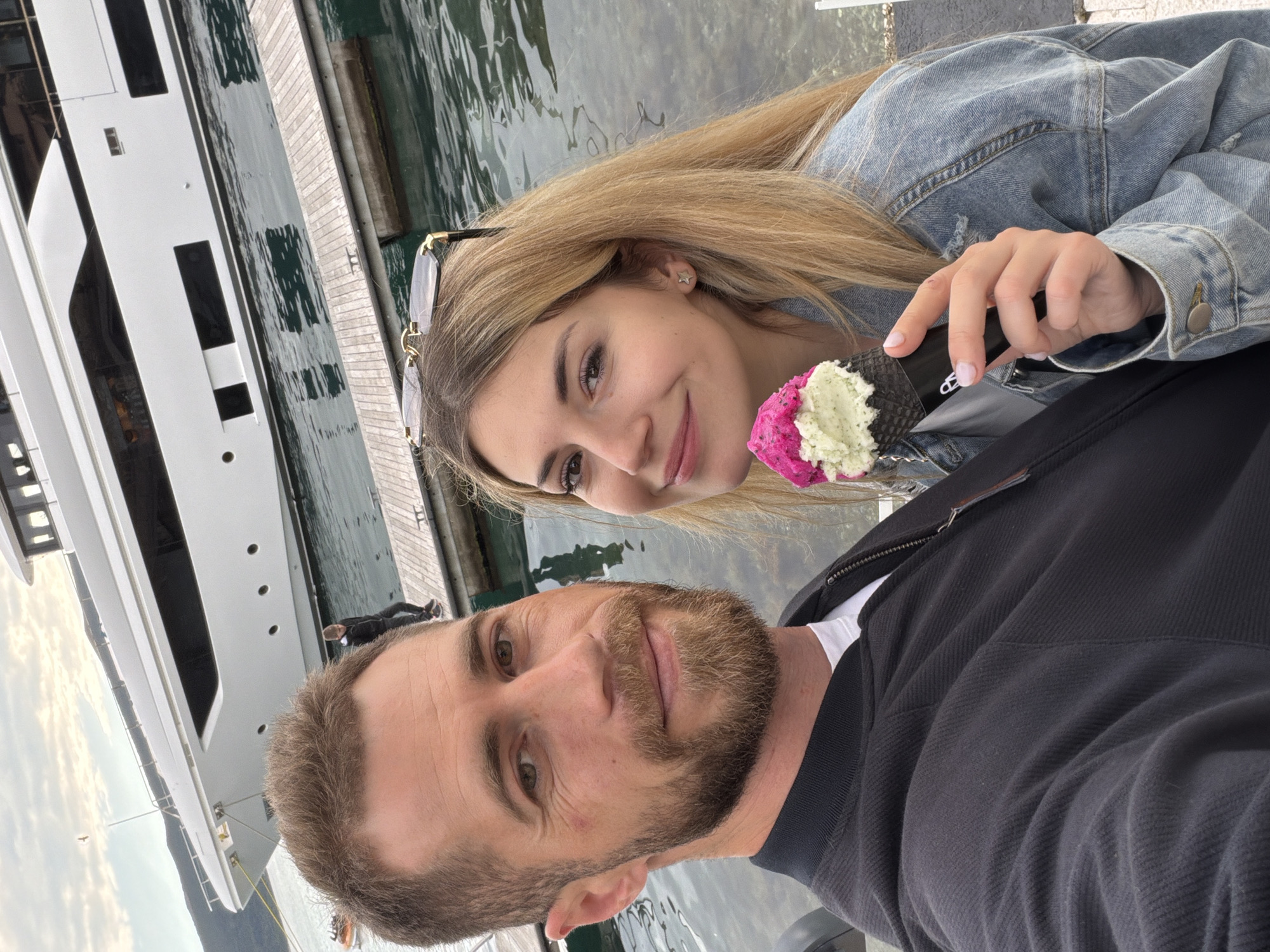 A selfie of a man and a woman, taken on a pier in Tivat, Montenegro. The woman is blonde and wears a denim jacket, holding a pink and white ice cream cone with a black waffle cone. The man has short brown hair and a beard and wears a black jacket over a white t-shirt. Behind them is a large white yacht docked at the pier. The water is visible, and the pier is made of wood. Sunglasses are perched on the woman's head.