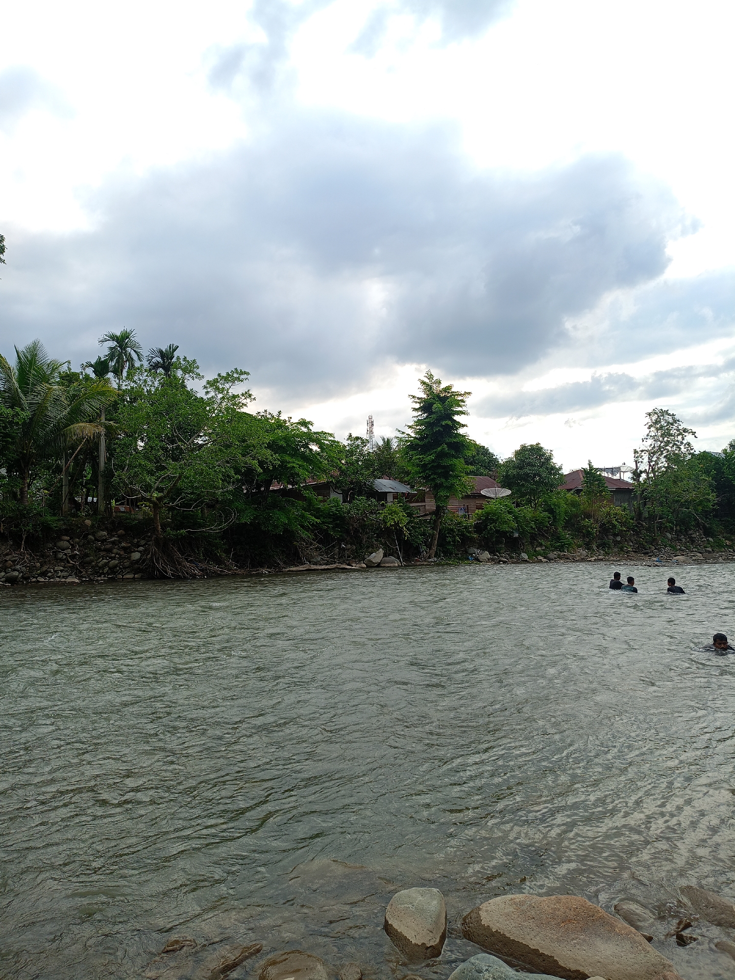 The image shows a river in Meurah, Indonesia, with several people swimming. The river is wide and the water is a murky green. There are trees and foliage lining the riverbank. The sky is overcast with heavy clouds. Rocks are visible on the foreground riverbank. A satellite dish and cell tower are visible amongst the trees.
