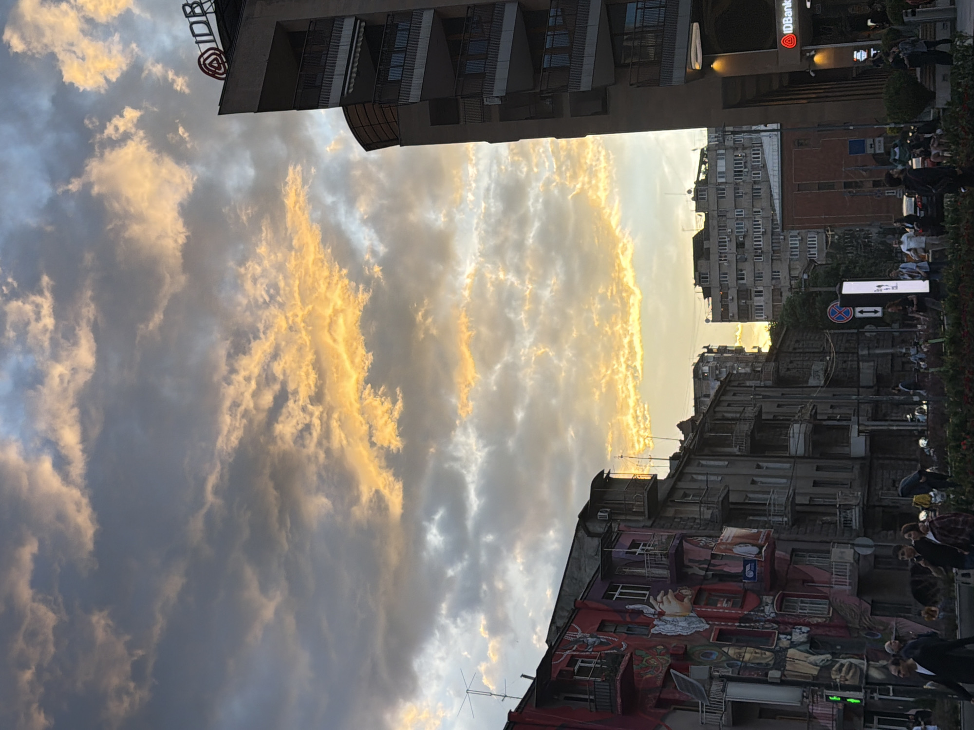 The image captures a street scene in Yerevan, Armenia, at dusk. The sky is filled with dramatic clouds illuminated by the setting sun, casting a golden light. Buildings of varying heights line both sides of the street.
On the right side of the street, there is a crowd of people, with some appearing to walk, while others seem to be standing or gathering near what looks like a street market or a pedestrian area.
On the left, buildings are diverse in their architectural styles. The nearest building has a visible mural. Higher up, the buildings are residential.
Signs are present on buildings, including the IDBank logo. A street sign with arrows is visible.