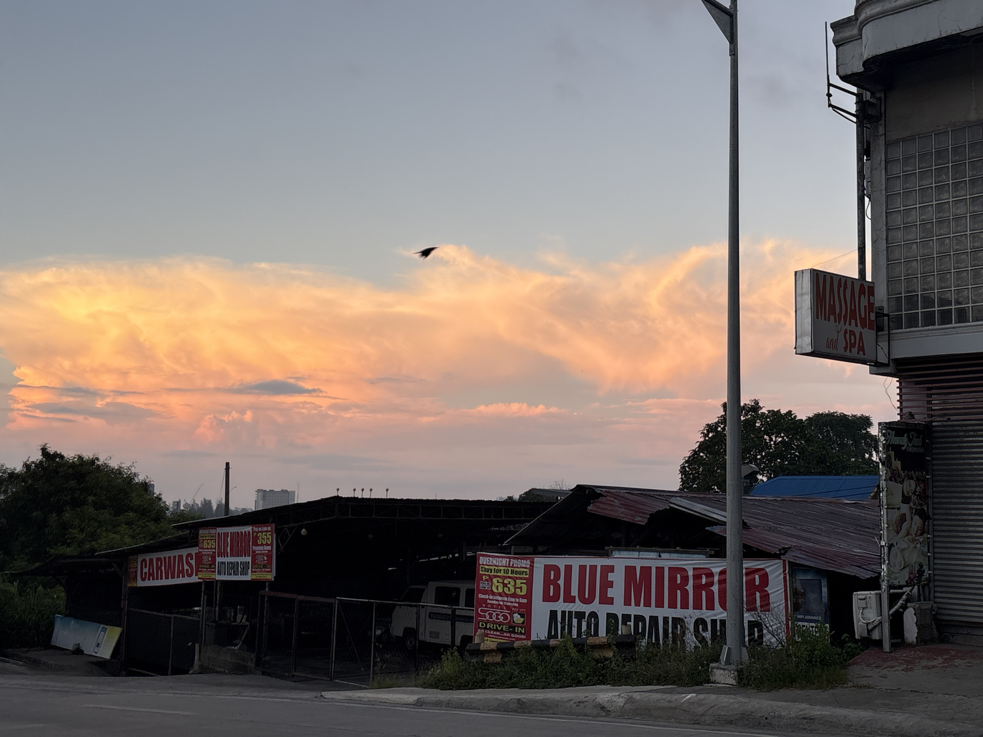 The image captures a street view in Davao City, Philippines, at dusk. The sky is a soft gradient of blue transitioning to warm orange hues from the setting sun illuminating the clouds. A lone bird is visible flying against the colorful sky. In the foreground, a row of low-rise structures line the street. On the left, a "CARWASH" sign is prominently displayed. Adjacent to it, a business with a sign advertising "BLUE MIRROR AUTO REPAIR SHOP" is situated. A white van is parked under the shelter of the auto repair shop. To the right, a building with a "MASSAGE and SPA" sign is partially visible, featuring a facade with tiled windows. A street light pole stands tall on the left side of the frame. The overall scene suggests a commercial area, likely a roadside establishment, at the end of the day.