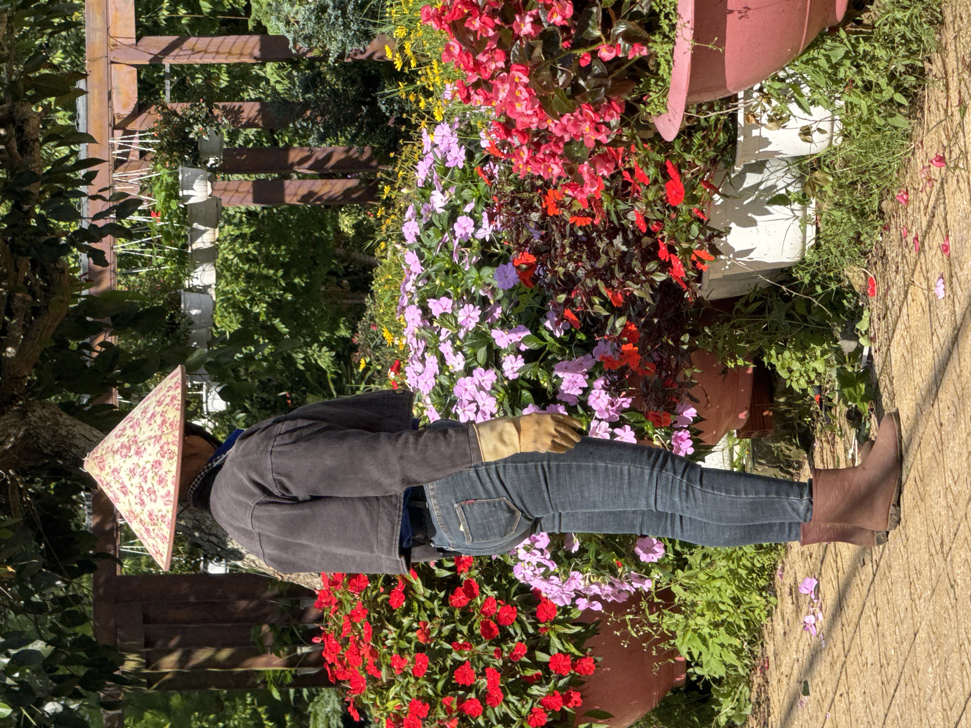 A person wearing a conical hat, a dark jacket, and jeans stands in a garden with a variety of flowers in pots. The person is wearing brown boots and yellow gloves. The garden appears to be well-maintained, with lush green foliage and colorful blooms. The background shows a wooden structure and more greenery. The lighting suggests it is daytime. The presence of the conical hat and the lush floral setting are characteristic of gardens and flower farms often found in and around Da Lat, Vietnam, a city known for its extensive flower cultivation and picturesque landscapes.