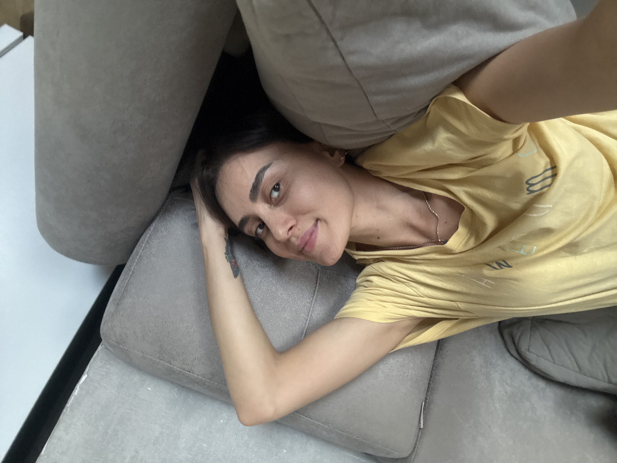 A young woman with dark hair and dark eyes is lying on a gray sofa, her head resting on a pillow. She is wearing a yellow t-shirt with some lettering on it. Her left arm is bent and resting behind her head, and her right arm is extended down her side. She is looking directly at the camera with a slight smile. The sofa appears to be in a modern living room. The background is blurred, but it seems to be a bright, neutral-colored wall. There is no indication of the specific location in Tbilisi, Georgia.