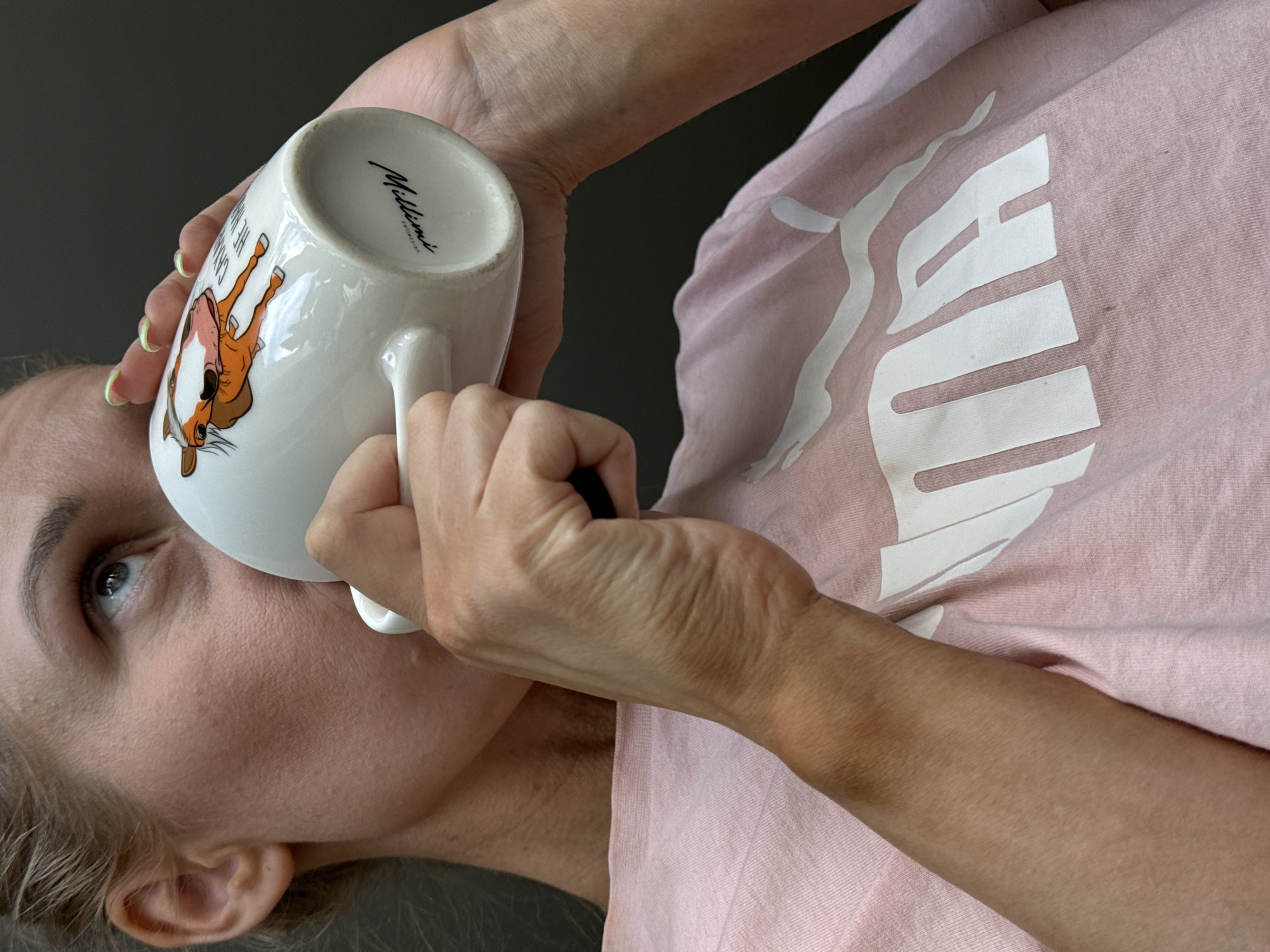 A person with fair skin and blond hair is holding a white ceramic mug with a horse illustration and the inscription "William's Collection." The person is drinking from the mug, their left eye visible. The person is wearing a light pink Puma t-shirt. The background is a plain, dark gray color. The location is not discernible in the image and could not be identified as being in Chaikovsky, Russia.