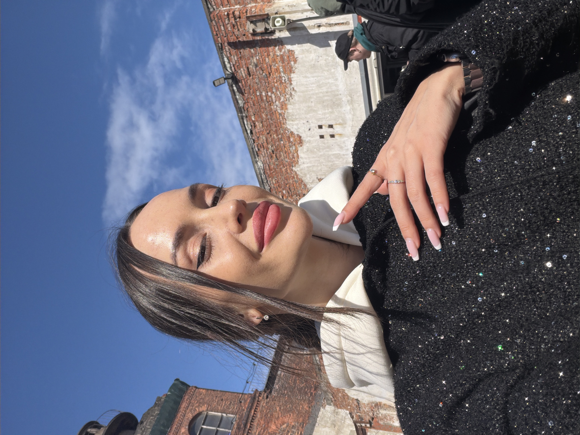 The image shows a woman with dark hair, light skin, and pink lips, facing up towards the camera. She is wearing a white hooded sweatshirt underneath a black jacket with sparkly accents. Her right hand is visible, adorned with rings, with long, well-manicured nails. In the background, a brick building with a window is visible, along with a clear blue sky with some clouds. A blurry figure of a person in a black jacket and a cap is partially visible in the upper right corner. The photo appears to be taken in an outdoor setting, potentially in the city of Saint Petersburg, Russia, based on the architectural style of the brick building.