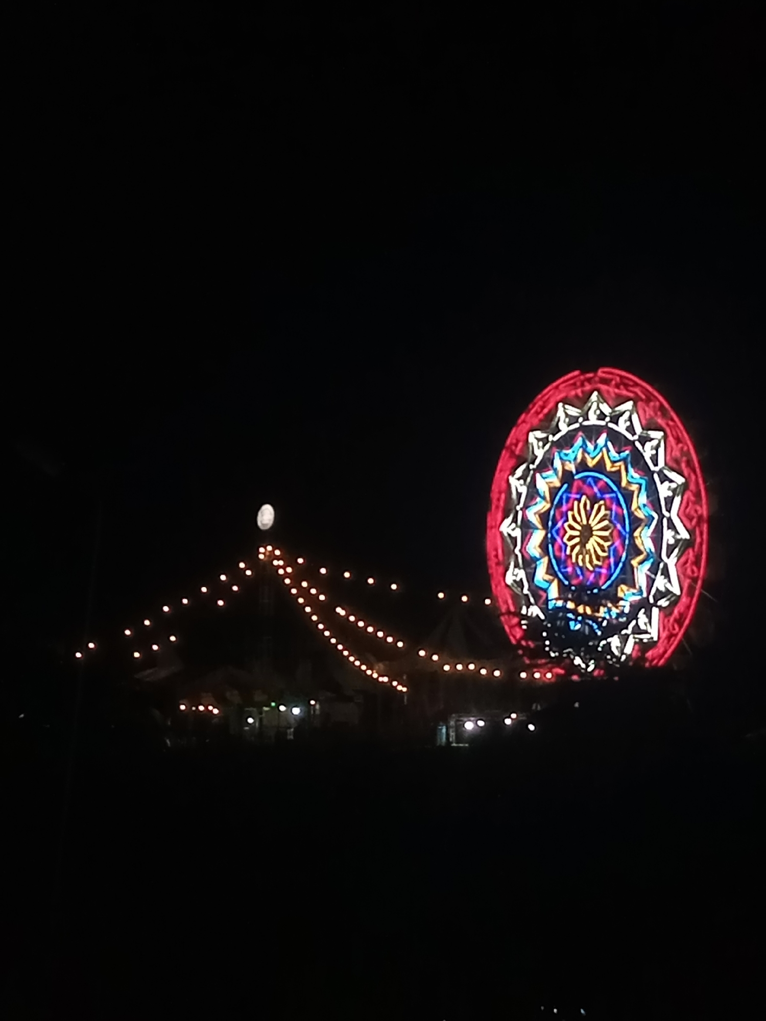 The image captures a night scene in Sigli, Indonesia, featuring a large, illuminated Ferris wheel decorated with a colorful, star-like pattern of lights. Strings of small, warm-toned lights stretch across the middle ground, creating a festive atmosphere. In the background, silhouettes of buildings and trees are visible against the dark sky, with a faint hint of what appears to be the moon or a bright light positioned above a central structure adorned with lights. The overall impression is of a nighttime festival or fairground.