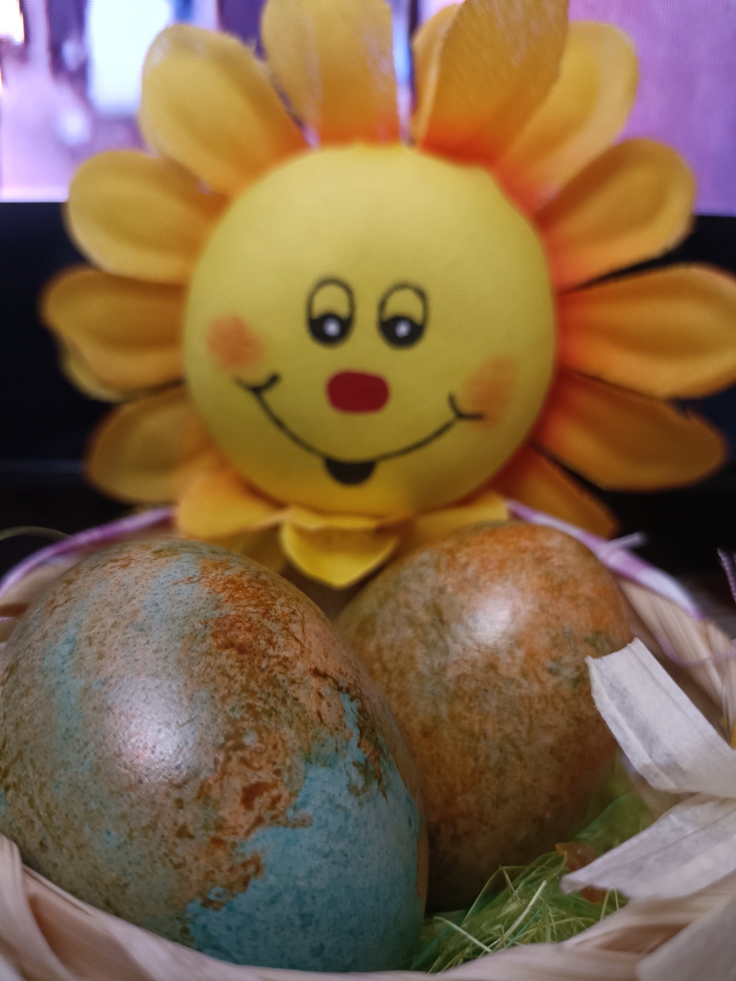 The image shows a close-up of two decorated Easter eggs in a woven basket. Behind the eggs is a stuffed, yellow sun-shaped toy with a smiling face. The eggs are colored with mottled patterns of blue, brown, and green. White flower petals and green decorative grass are visible in the basket. The background is blurry, but suggests an indoor setting. The image does not provide any information to determine a location in Plovdiv, Bulgaria.