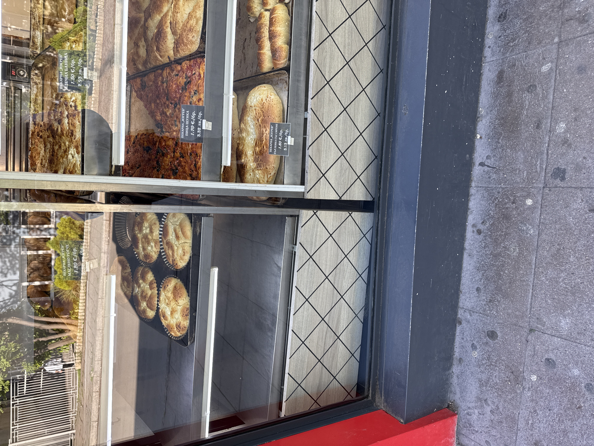 The image shows a window display of a bakery, featuring various baked goods. The bakery is called "Изгрев" (Izgrev). The window contains several shelves with different types of bread, pastries, and pies, each with a price tag in both euros and Bulgarian leva. The baked goods visible in the window include milk pastry (Млечна Баница), ham pizza (Пица Шунка), and cheese pie (Пърленка Със Сирене). A price sign also indicates a butter croissant for 1.20 EUR or 2.34 BGN. The window also reflects the outside environment, showing trees, buildings, and a white metal bench. The window frame is dark grey with a red base. The sidewalk is made of grey stone tiles.