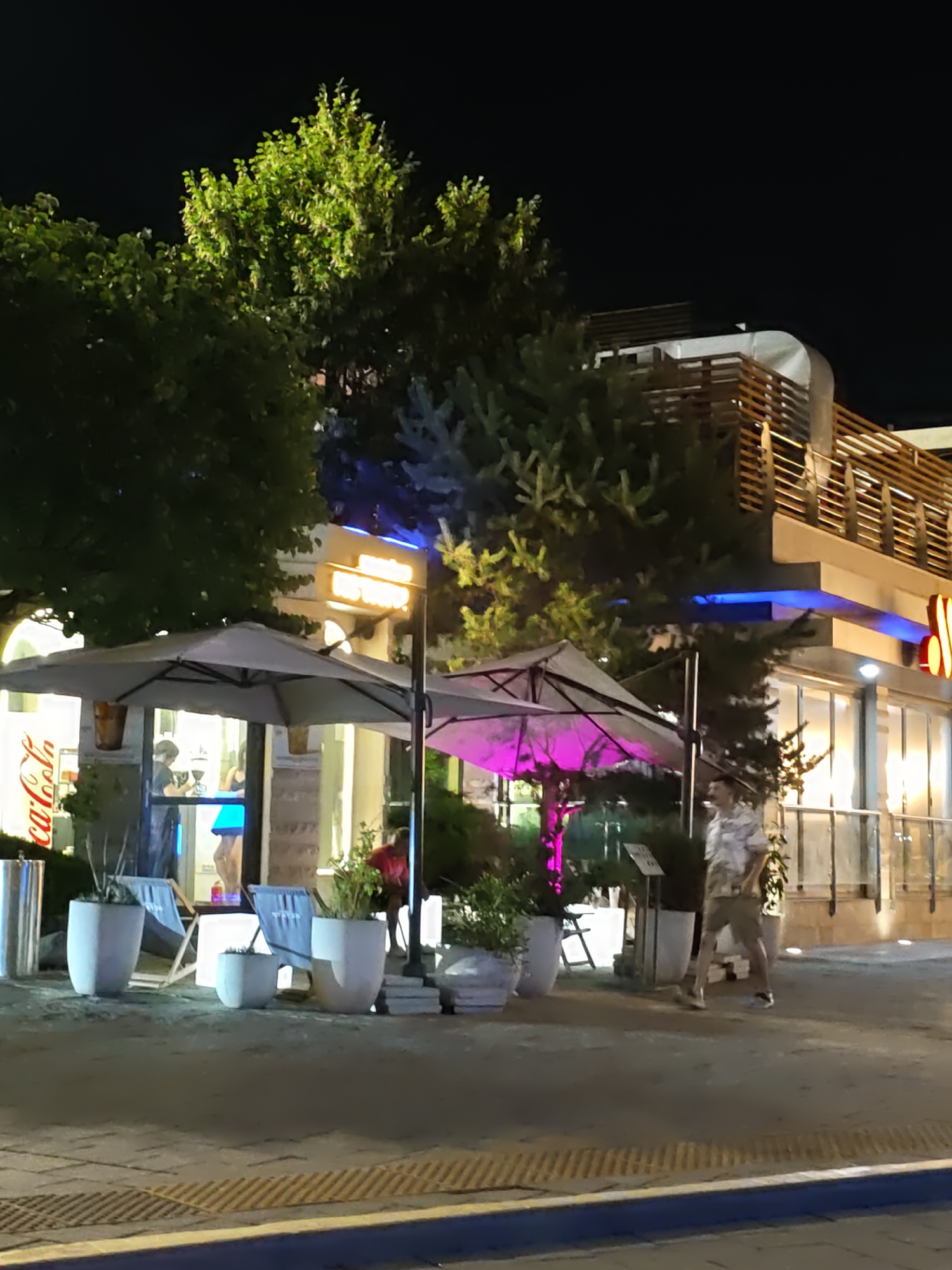 The image depicts a street scene at night, likely in Rostov-on-Don, Russia, outside a commercial establishment. A man in a patterned shirt and shorts is walking on the paved sidewalk. To his left, there are large potted plants and outdoor seating areas with umbrellas, one of which is illuminated with purple light. Behind the seating area is the building's facade, featuring large windows and a sign with illuminated lettering. Trees and foliage are visible, some illuminated by artificial lighting. The overall ambiance suggests an outdoor dining or recreational area during the evening.
