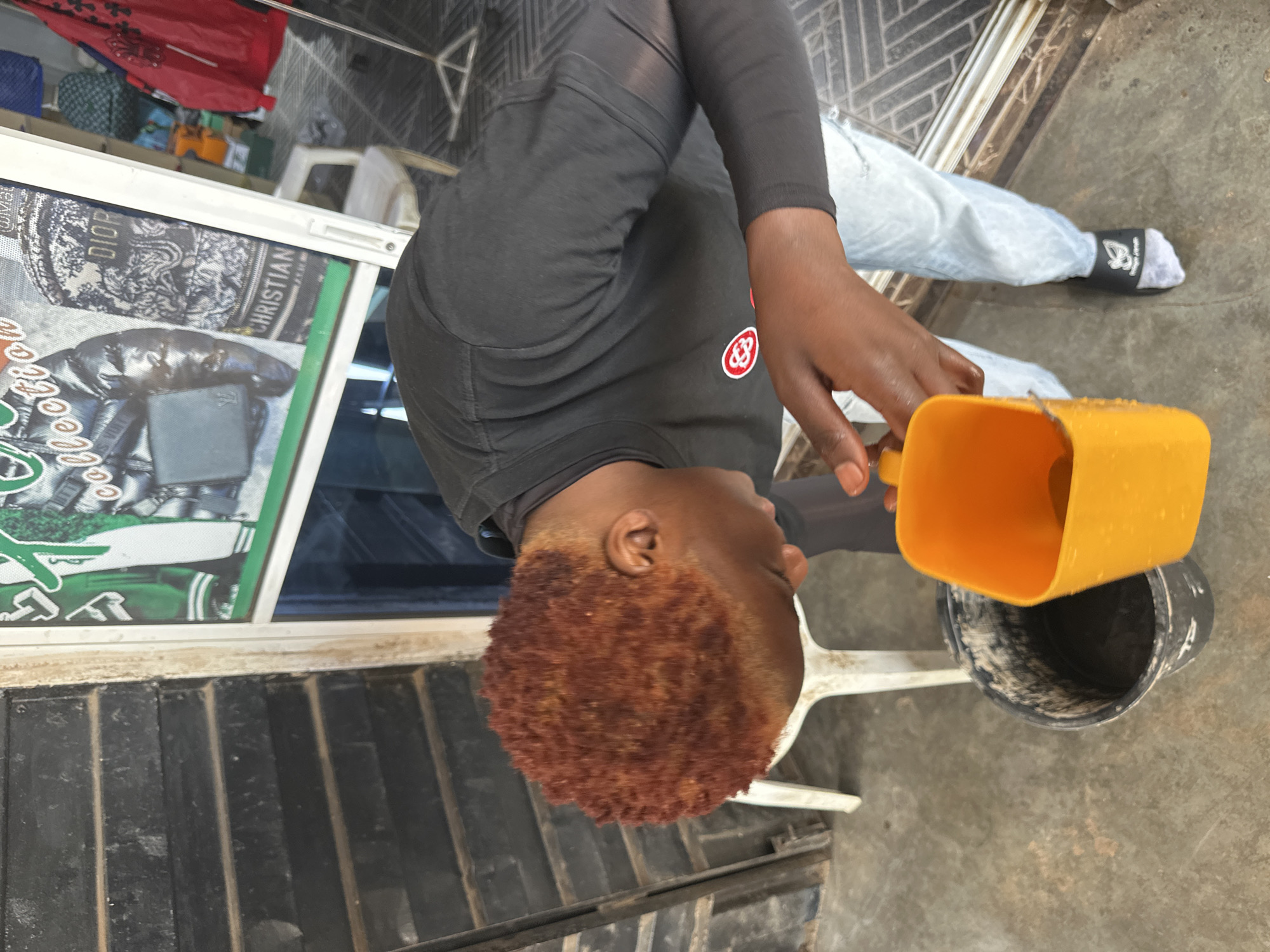 A young person with reddish-brown, short, textured hair is bending over a black bucket filled with a dark liquid. They are holding a bright orange plastic scoop, tilted as if to pour liquid from it into the bucket. The person is wearing a black long-sleeved shirt with a circular red and white logo on the chest and light-wash jeans. They are also wearing one white sock with a black band that has a white logo on their left foot. The scene appears to be outdoors, with a dark, slatted wall to the left and a shop window displaying clothing with brand names like "DIOR" and "CHRISTIAN" to the right. The ground is concrete. The image was taken in Bukuru, Nigeria.