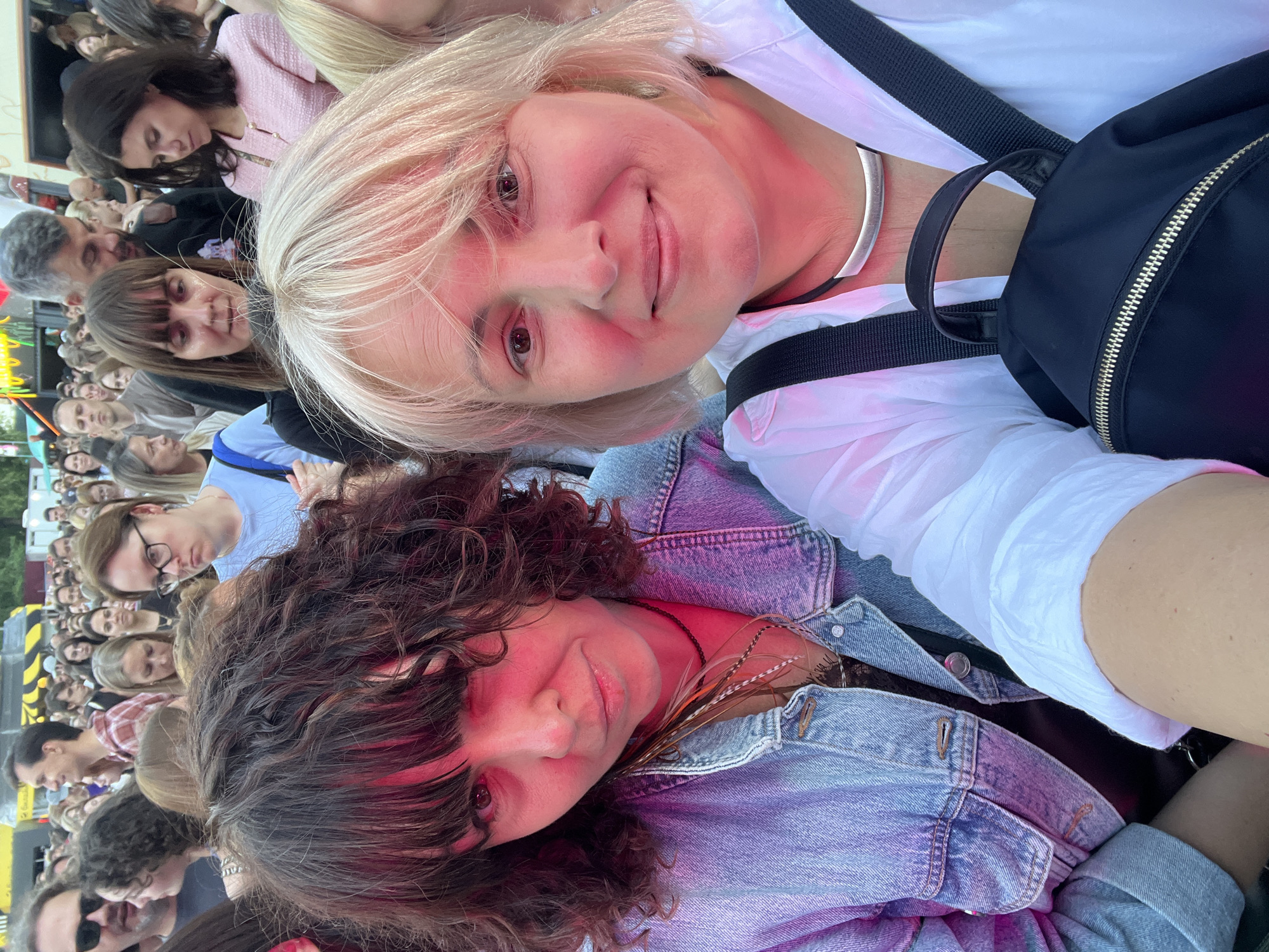 Two women, one with dark curly hair and the other with blonde hair, are in the foreground of this image, smiling at the camera. They are at an outdoor event, likely a concert or festival, with a crowd of people visible in the background. The blonde woman is wearing a white collared shirt and a black backpack, while the woman with dark hair is wearing a denim jacket. The lighting suggests it is daytime or early evening, with some artificial lighting adding color to the scene. The general environment appears to be an urban or suburban setting in Khimki, Russia, given the context provided.