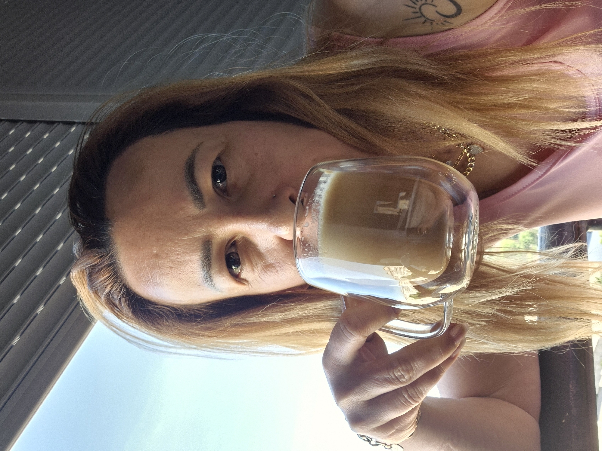 A medium shot shows a person with light brown hair holding a clear glass mug of coffee. The person is looking towards the camera. Their face is partially obscured by the mug and their hair. They are wearing a pink tank top and a necklace with a heart-shaped charm. A tattoo of a sun is visible on their right shoulder. The background shows a blurred view of a building and a light blue sky, suggesting an outdoor setting. The lighting is natural and bright, indicating it is daytime.