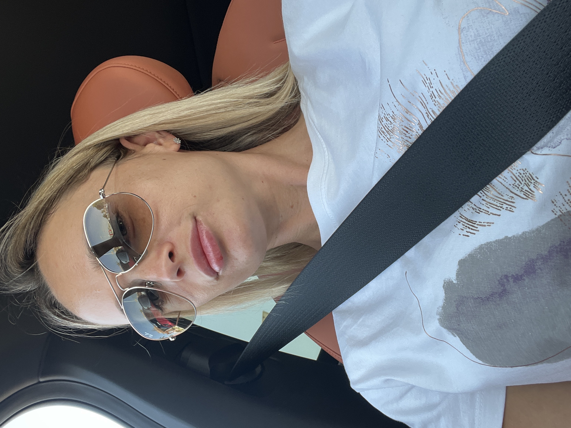 A selfie of a woman wearing sunglasses and a white t-shirt is taken inside a car. She has blonde hair and is wearing a silver necklace. The car has a brown leather seat and a black seatbelt is visible across her chest. The background shows a window and a glimpse of a blurry outdoor scene. The image is shot from a high angle, looking down at the woman.