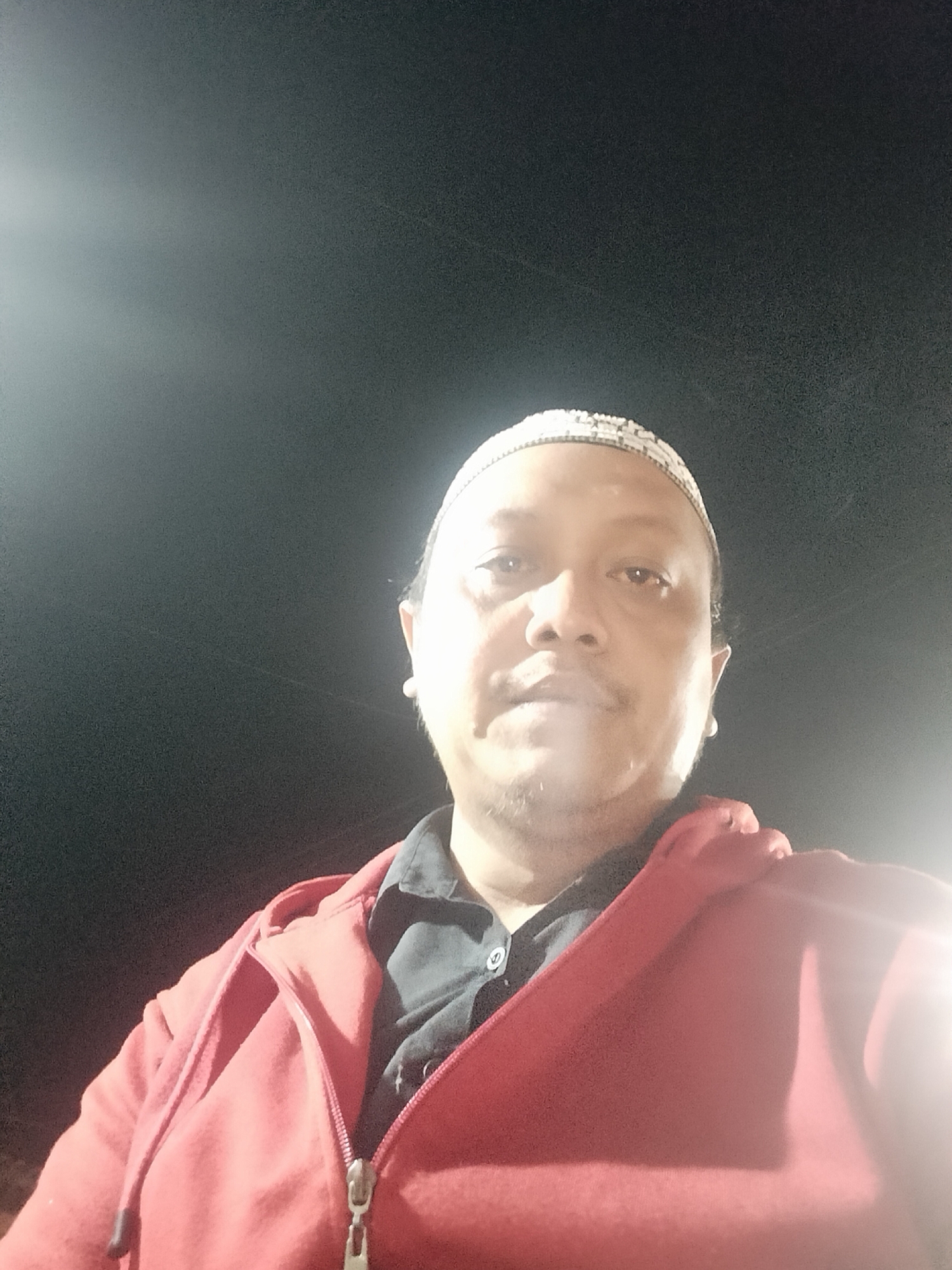 A low-angle, close-up selfie shows a person wearing a red zip-up hoodie over a dark collared shirt. They are also wearing a white, embroidered skullcap. The background is dark and appears to be outdoors at night, with a bright light source causing lens flare in the upper left and lower right of the image. The person has dark hair, a mustache, and light-colored earrings. The scene is likely in Srono, Indonesia, given the metadata.