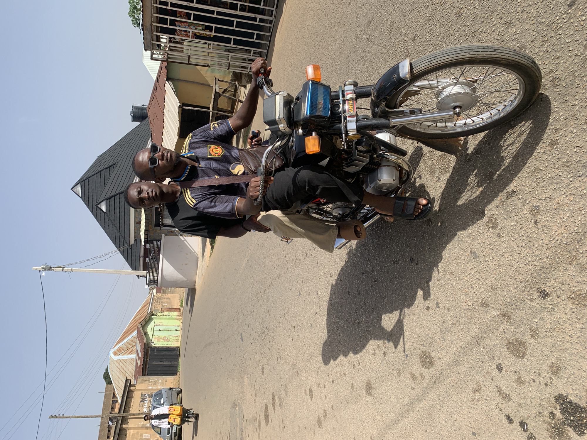 Two men are riding a black motorcycle on an asphalt street in Jos, Nigeria. The rider wears a black and yellow shirt, black pants, and sandals, and has sunglasses on. The passenger is in front of him wearing a black shirt and khaki pants. The motorcycle is pointed toward the bottom of the frame. Street scenes with buildings are visible in the background, with power lines running overhead. Another vehicle, carrying water containers on its roof, is further down the street. The sky is light blue. The motorcycle's shadow is cast on the road.