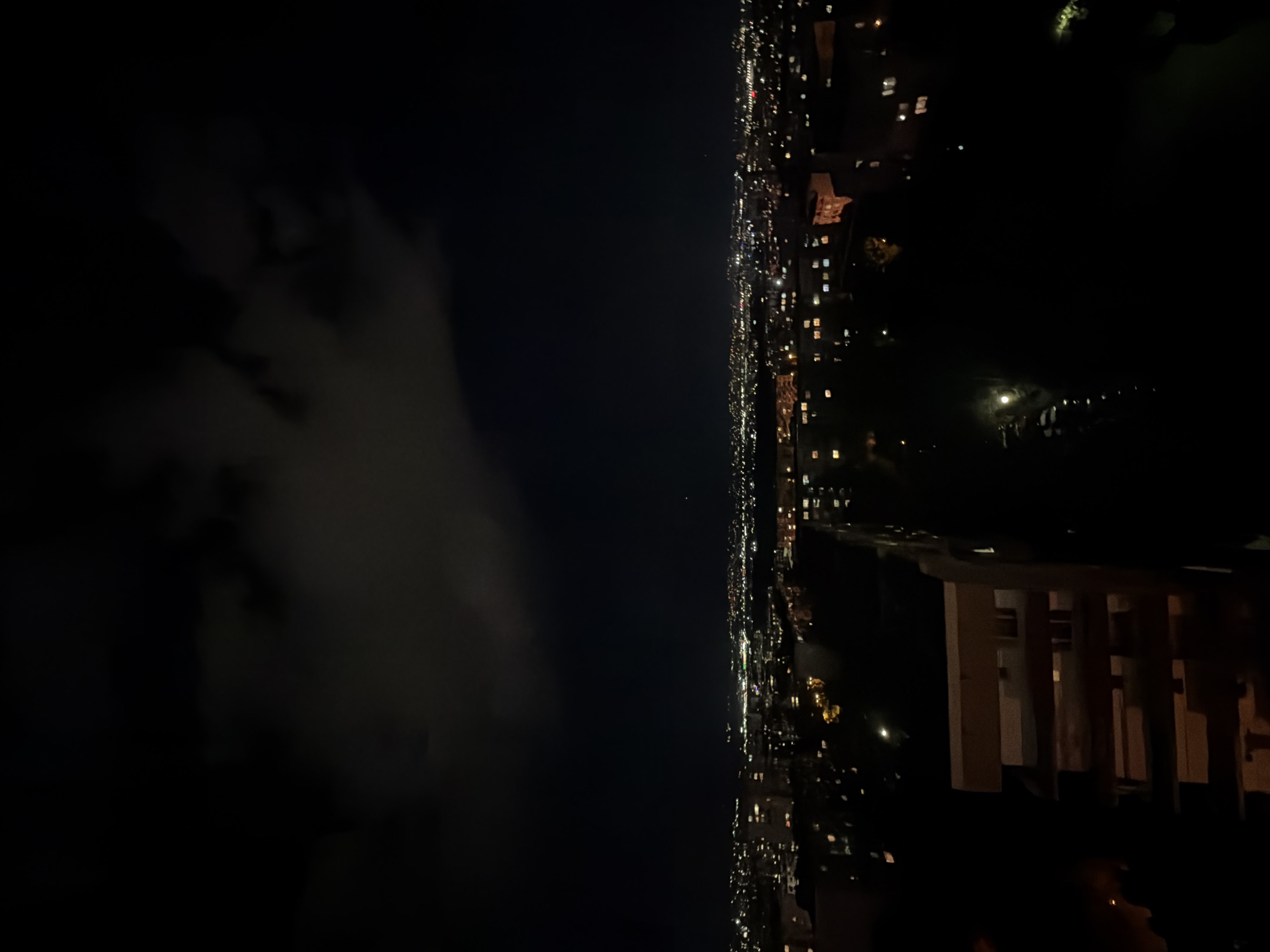 The image captures a vertical, nighttime view of Sofia, Bulgaria, depicting a sprawling urban landscape illuminated by artificial lights. The right side of the frame is dominated by the cityscape, where countless lights from buildings and streets create a vibrant, glowing line that extends from the bottom to the top, signifying a vast urban expanse. Numerous windows glow from the dark facades of buildings. The left side of the image is largely filled with the dark night sky, where faint, wispy cloud formations are visible, adding subtle detail to the darkness. In the lower right foreground, a part of a building or structure with distinct architectural elements, possibly a balcony railing, is dimly visible, indicating the vantage point. The time of day is clearly night, and the weather appears to be partially cloudy. There are no people or specific activities discernible within the frame, and no visible text.