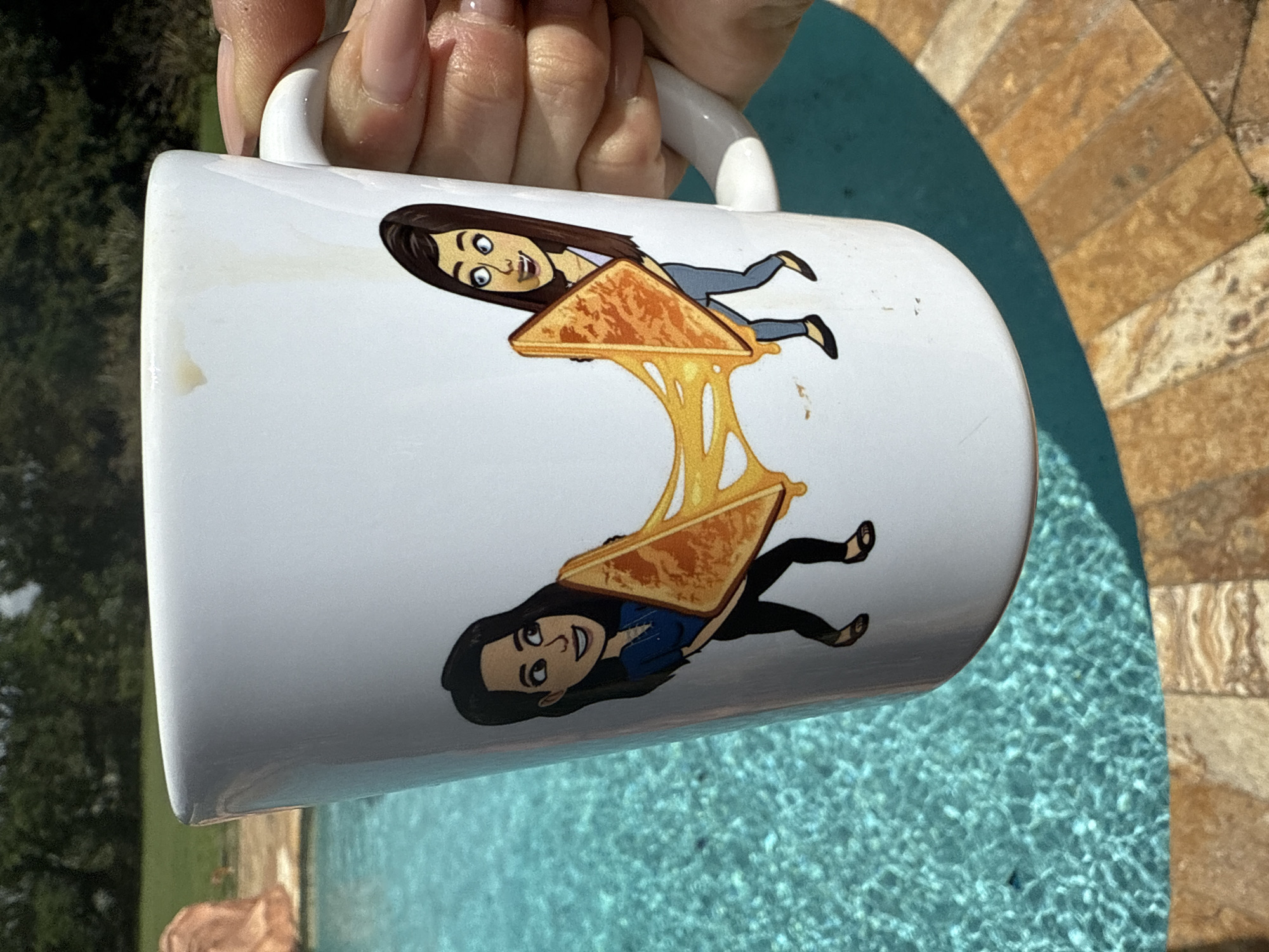 Here's a concise, forensic description of the image:
A white ceramic mug is held by a fair-skinned hand with manicured nails. The mug features two cartoon images of the same person, each seemingly held up by a grilled cheese sandwich with visible melted cheese strings connecting them. The background includes what appears to be a swimming pool with textured tiles or coping. Part of the location appears to have vegetation and trees. This scene is likely taken in Davie, United States of America.