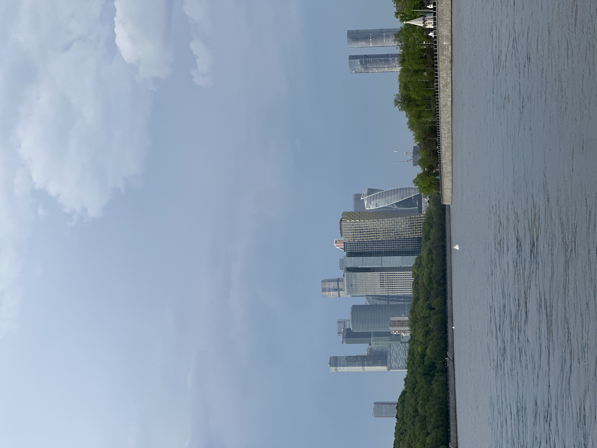 The image depicts the Moscow International Business Center (Moscow-City) skyline from across the Moscow River. The river is visible on the right side, with a concrete embankment running along the edge where trees line the bank. The sky is partially cloudy. The Moscow-City skyscrapers are visible in the distance. There are no people visible in the image. A small boat with a white sail is visible in the Moscow River.