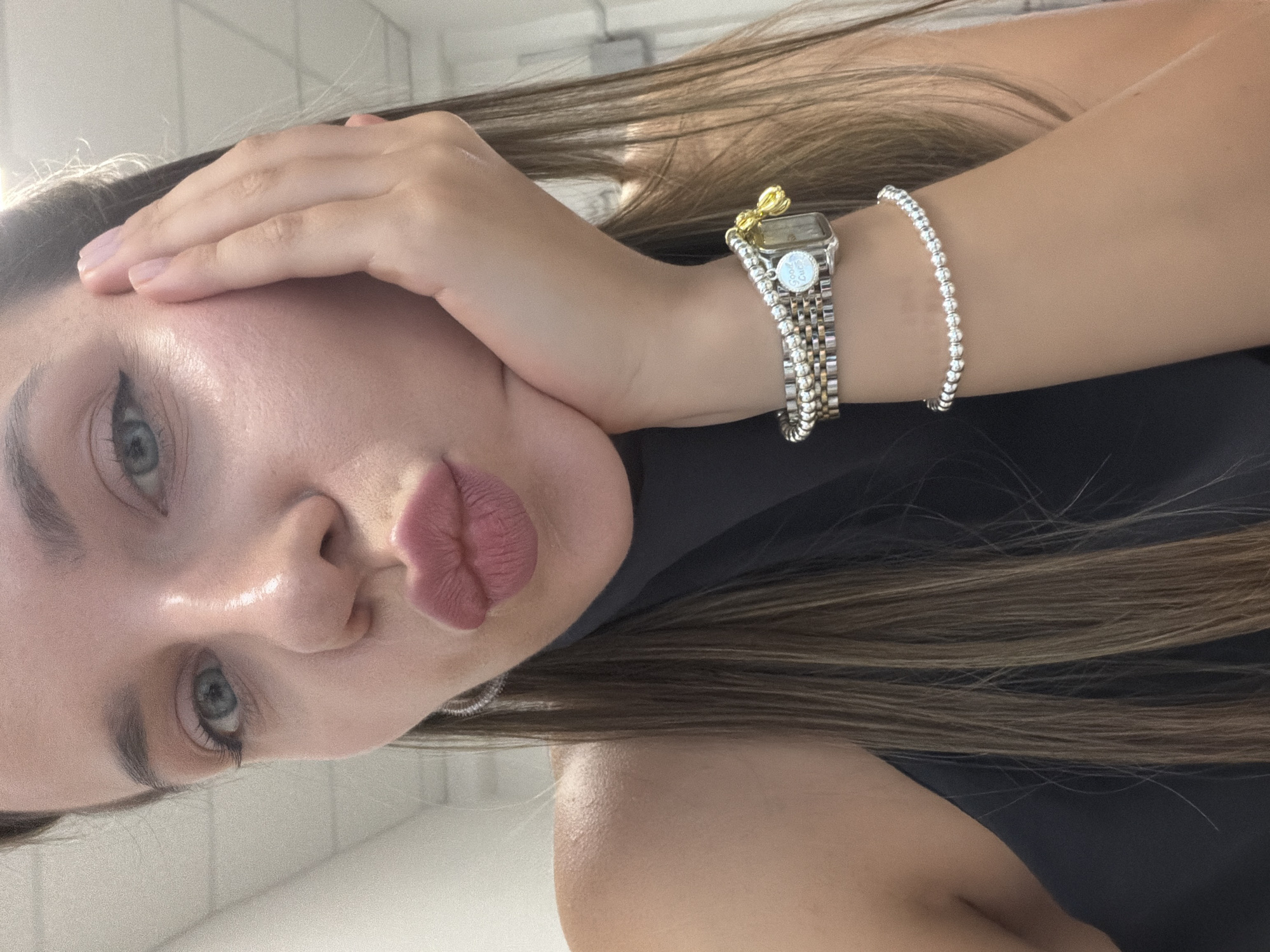 The image shows a close-up, self-taken portrait of a woman with blue eyes and full lips pursed in a kissing gesture. Her right hand is supporting her face, and she is wearing several bracelets on her left wrist. The background appears to be an indoor setting with light-colored walls and a ceiling that suggests an office or similar environment. The woman has long, brown hair with blonde highlights, and is wearing a dark-colored top. There is no discernible indication of the location being Homyel, Belarus.