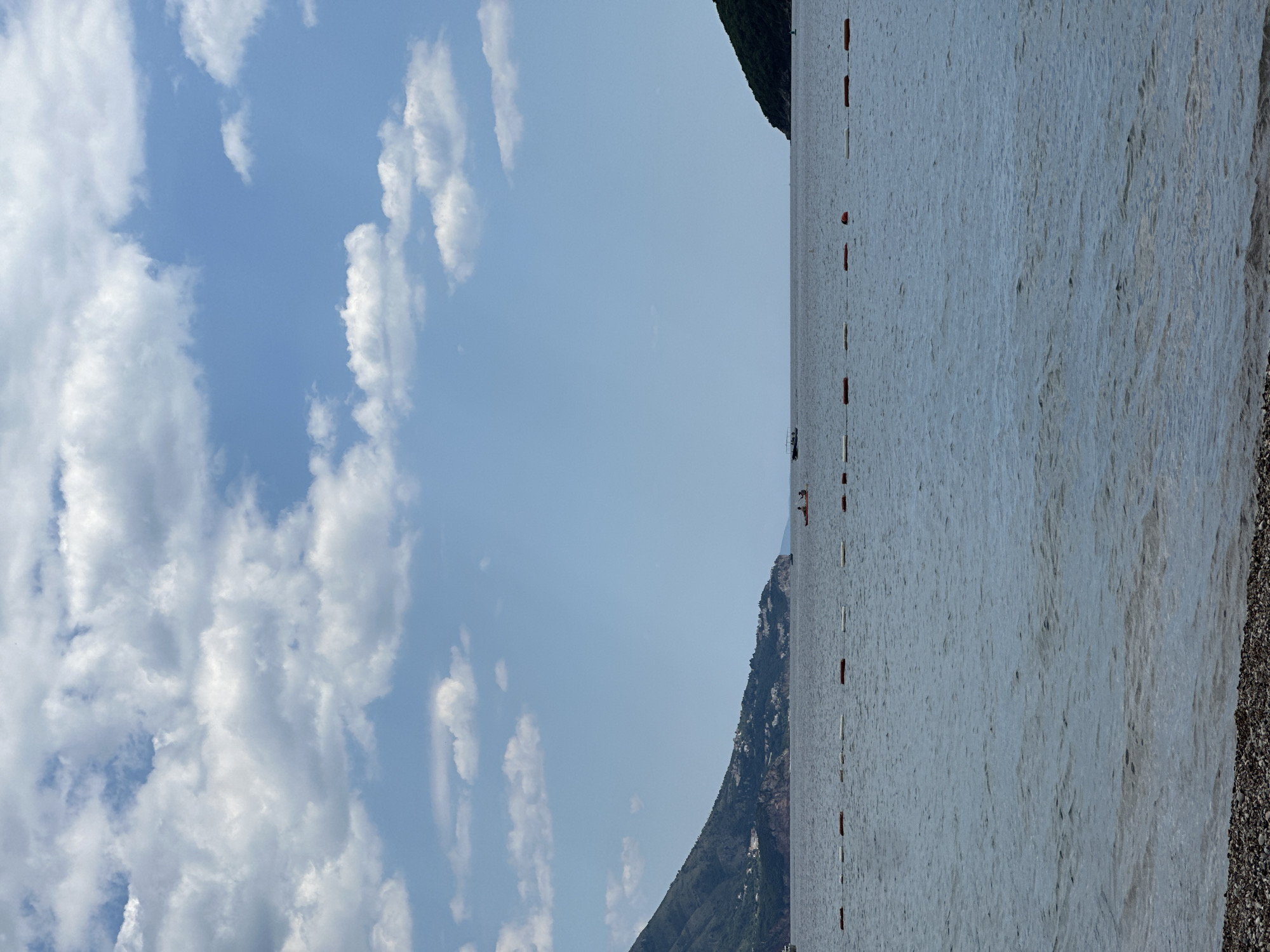The image shows a beach scene in Budva, Montenegro. The left side is dominated by a blue sky with scattered white clouds. The right side features the Adriatic Sea, appearing calm with a grayish hue. A safety line marked by intermittent orange floats stretches across the water, delineating a swimming area. In the distance, a mountainous backdrop is visible, partially obscured by haze. The foreground consists of a pebbly beach. No distinct human activities or interactions are evident within the frame.
