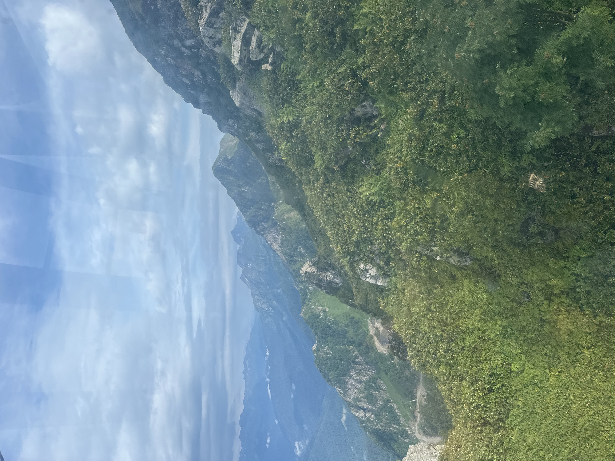 The image shows a panoramic view of the Caucasus Mountains near Estosadok, Russia. In the foreground, lush green vegetation, including trees and bushes, covers the steep mountain slopes. Rocky outcrops are visible, interspersed with patches of green. In the middle ground, more mountains are visible, with a road winding its way up one of the slopes. The background features a series of mountain ranges fading into a hazy blue distance under a cloudy sky. The scene appears to be captured from a high vantage point, possibly from a cable car or a mountaintop. There are no people or animals visible in the image. The overall impression is one of natural beauty and vastness.