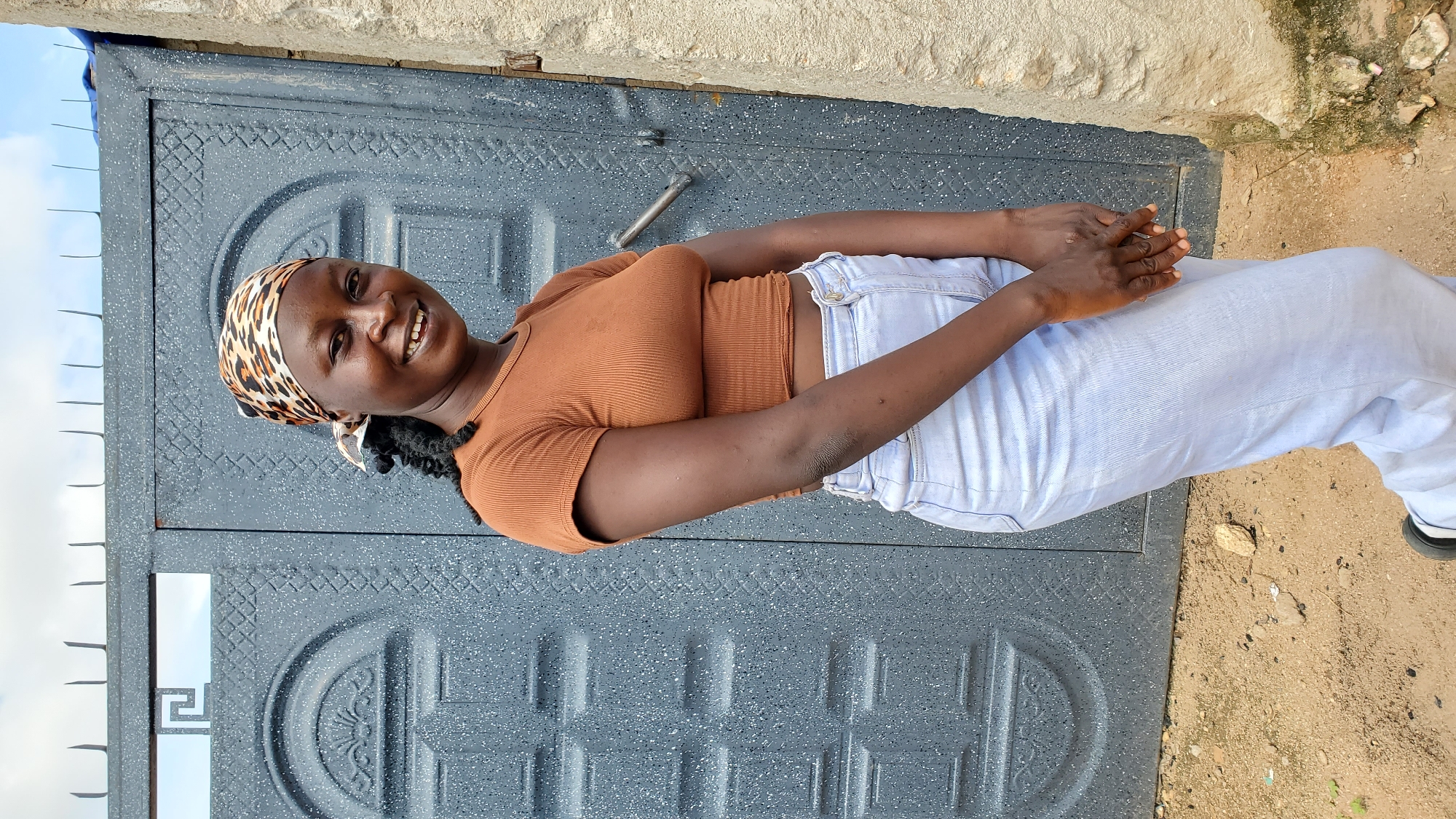 A Black woman wearing a brown, short-sleeved, ribbed crop top and light blue denim pants stands in front of a large, dark gray metal gate with ornate carvings. She has a leopard-print headscarf tied around her head, and her dark hair is braided. She is smiling and looking at the camera. Her hands are clasped in front of her. The gate appears to be a double door with a textured surface and a horizontal handle on the right side. The ground in front of the gate is sandy and contains small stones. To the right of the gate is a rough, beige-colored concrete wall. The sky above is partly cloudy. The overall scene suggests an outdoor setting, possibly a residential area in Jalingo, Nigeria, given the architectural style of the gate and the prevalence of this type of wall construction in the region.