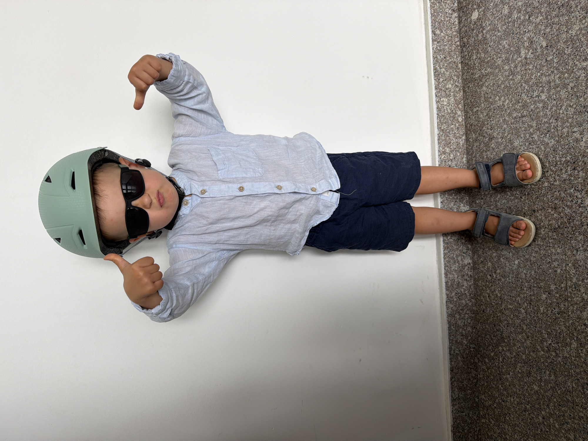 A child wearing a light blue button-down shirt, navy blue shorts, grey sandals, a light green helmet, and black sunglasses lies on their back with their left thumb pointed down and right thumb pointed up. The child is positioned between a white wall and a speckled brown and grey tile floor. The location is likely indoors, but the context of the city of Budva, Montenegro, is not visually apparent.