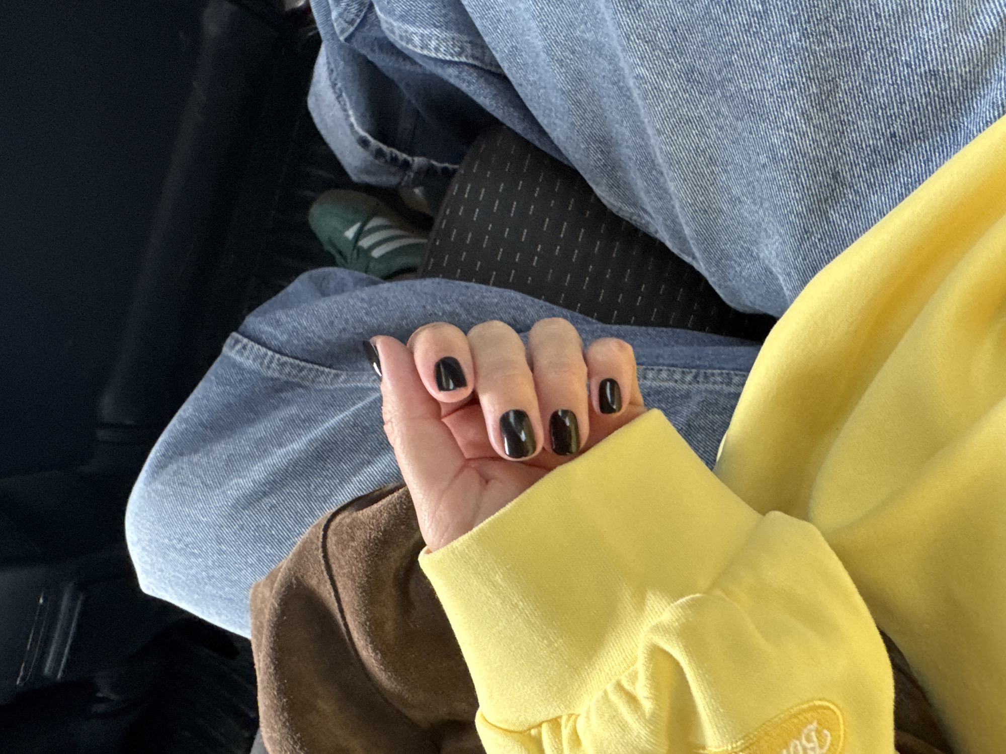 This image, captured in Chaikovsky, Russia, offers a close-up perspective of a person's hand and lower body, seen from above. The focal point is a hand with a light skin tone, adorned with glossy black nail polish on all visible fingernails.
The individual is dressed in casual attire. A bright yellow long-sleeved top, possibly a sweatshirt or hoodie, is visible, featuring a soft, somewhat plush texture. Near the cuff, there is faint yellow embroidery, appearing to spell out a word like "Butter." Below the hand, a section of a brown sleeve or fabric is seen on the left. The person's legs are covered by blue denim jeans, characterized by a distinct vertical weave pattern, suggesting a classic denim texture.
Peeking from beneath the jeans is a dark, patterned fabric, likely leggings or another layer, featuring small, vertical white dash marks. Further down, a dark green sneaker with prominent white stripes on the side is visible, suggesting an athletic shoe. The overall background is dark and indistinct, hinting at an indoor setting, possibly the interior of a vehicle such as a car or bus, where the person is seated. The image captures a quiet, seated moment, emphasizing the clothing and nail details. The time of day and specific weather conditions are not discernible from the photograph.