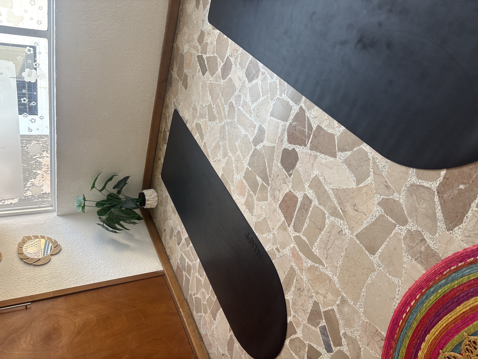 The image depicts a section of an interior space, with a focus on the textured floor and nearby wall. The floor is composed of irregular beige and brown tiles, creating a mosaic-like pattern. Resting on the floor are two elongated black mats, one positioned vertically and the other diagonally. The word "SHAPE" is visible on the lower mat. A portion of a colorful woven circular object appears in the lower right corner.
The wall is painted white, and a framed window is visible on the left. A green potted plant sits on a wooden ledge, adjacent to a heart-shaped decorative object hanging on the wall. The space appears to be within a building in Puebla, Mexico. No people are visible in the image.