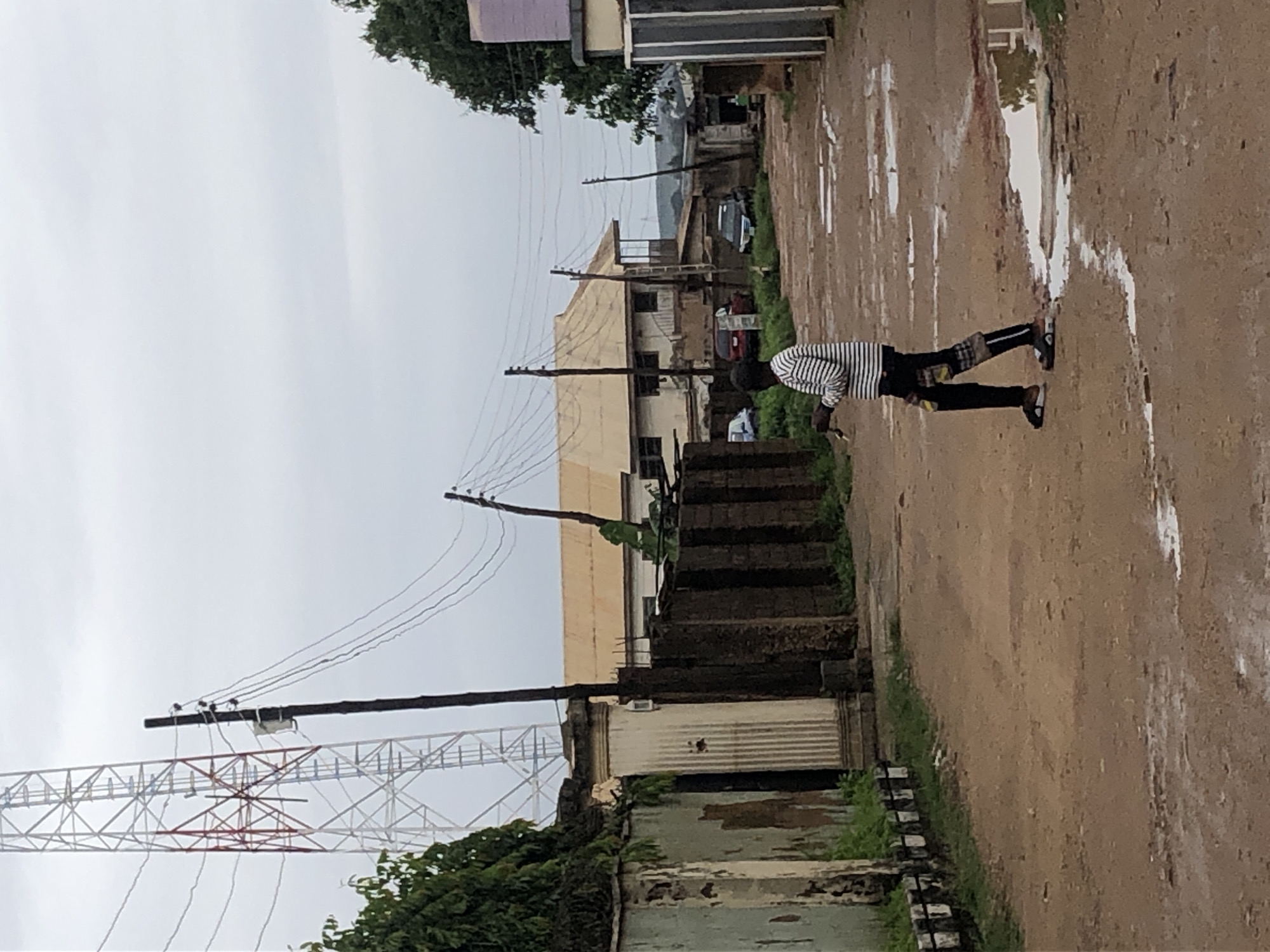 A young boy walks on a dirt road with puddles reflecting the overcast sky. He is wearing a horizontally striped shirt and black pants with patterned patches on the lower legs. In the background, a tall communication tower stands prominently, with utility poles and power lines crisscrossing overhead. The road is lined with various structures, including a dilapidated wall with green foliage, a building with a light-colored roof, and a brick wall. Several vehicles are parked along the road. The overall scene suggests an urban or semi-urban environment.