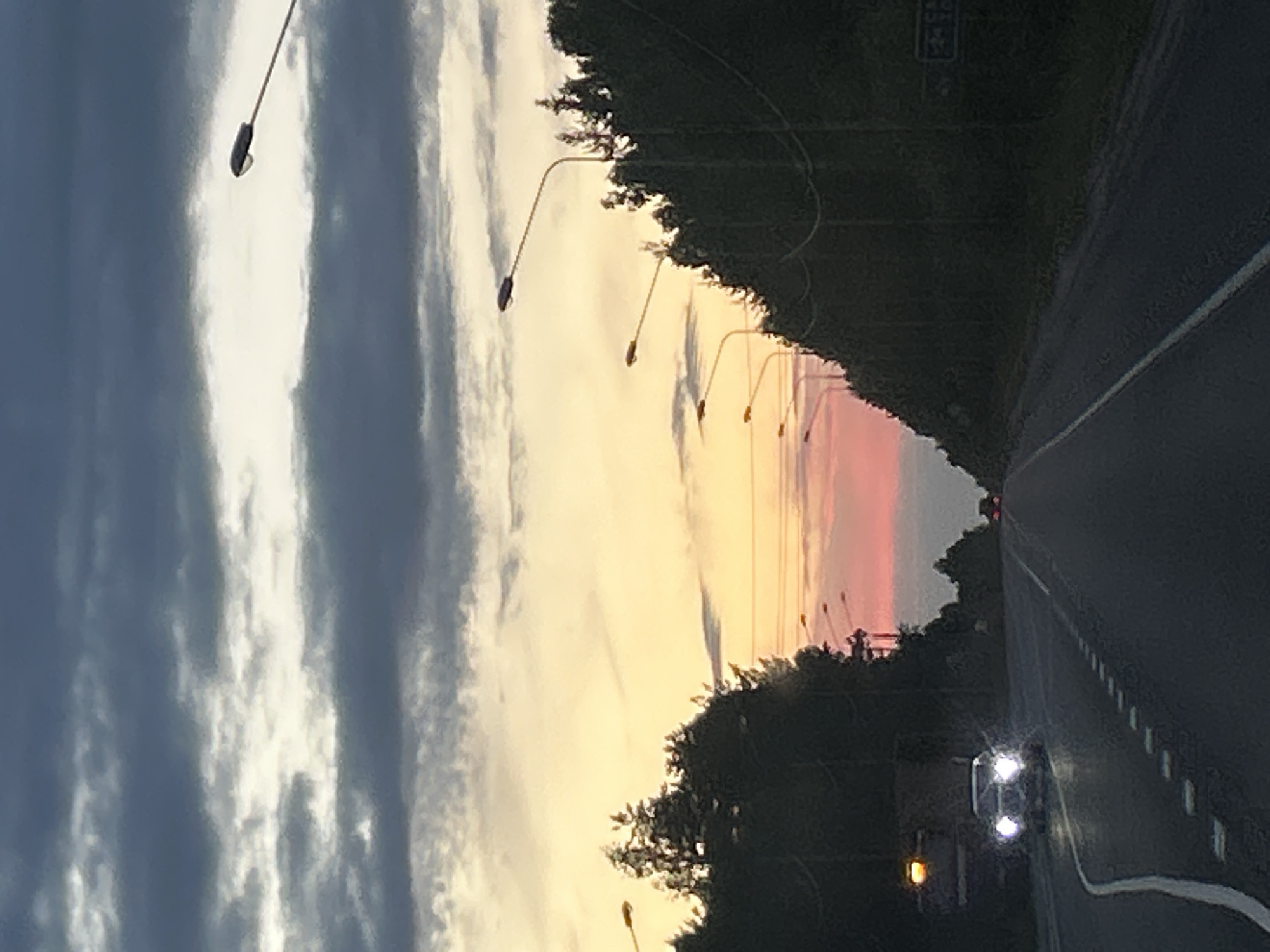 The image depicts a road scene at dusk, likely in Bel'skoe, Russia, with a dramatic sky transitioning from sunset colors to dark clouds. In the foreground, a dark asphalt road stretches into the distance, bordered by trees and a line of streetlights on the right. A white line marks the edge of the road on the left. A car with its headlights on is visible in the left lane, approaching the viewer. Another vehicle is further down the road. The sky above is filled with layered clouds, with a bright, yellowish-orange hue near the horizon, fading into pink and then dark grey clouds at the top. The overall impression is one of twilight on a rural or suburban highway.