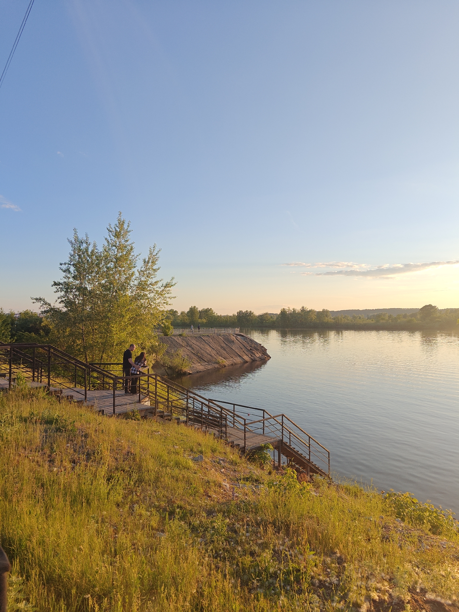 This image, captured in Nikolo-Beryozovka, Russia, depicts a serene riverside landscape under a clear blue sky transitioning to a warm, golden hue near the horizon, indicative of late afternoon or sunset. In the foreground, a grassy embankment covered in tall, dry-looking vegetation slopes downward. A multi-tiered wooden staircase with dark metal railings descends from the top of the embankment towards the river. Two individuals, one male in a dark shirt and one female in a plaid shirt, stand on an upper landing of these stairs, leaning against the railing and looking towards the water. Behind them, a large deciduous tree with bright green leaves is bathed in golden sunlight. The wide, calm river occupies the mid-ground, reflecting the sky and surrounding light, with gentle ripples visible on its surface. On the opposite bank, a treeline extends along the shore, with a distinctive sandy or earthen landform protruding into the water on the left. Several indistinct figures are visible on a raised path or structure along the far bank. White, fluffy detritus is scattered on the ground near the bottom right of the foreground.