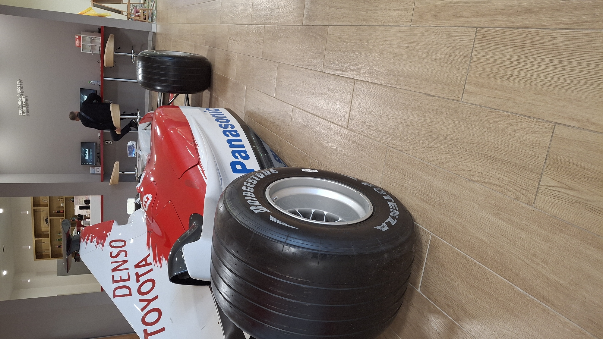 In Novogorelovo, Russia, a section of a red and white Formula 1 racing car is prominently displayed on a light brown tiled floor. The car features prominent "DENSO" and "TOYOTA" branding in red lettering on its white bodywork, and "Panasonic" in blue on its red side. A large, black racing tire with "BRIDGESTONE POTENZA" branding is visible in the foreground, mounted on a silver rim. In the background, a person wearing a black jacket is seated at a red counter, using one of two computer stations. The stations are equipped with monitors displaying the "ictv.ru" logo. The wall behind the counter has text that reads "СВОБОДНЫЙ ДОСТУП В ИНТЕРНЕТ ДОСТУП В ИНТЕРНЕТ" which translates to "FREE ACCESS TO THE INTERNET ACCESS TO THE INTERNET". The environment appears to be an indoor public space, possibly a showroom or a public internet access point.