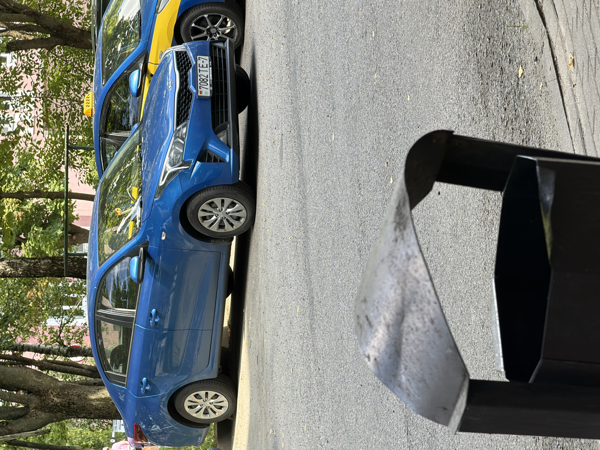 A blue Kia sedan with license plate 7082 TE-7 is parked on a street in Minsk, Belarus, next to a yellow and blue taxi. A dark metal trash can is in the foreground, partially obscuring the view of the cars. Trees and a building are visible in the background. The sky is overcast.