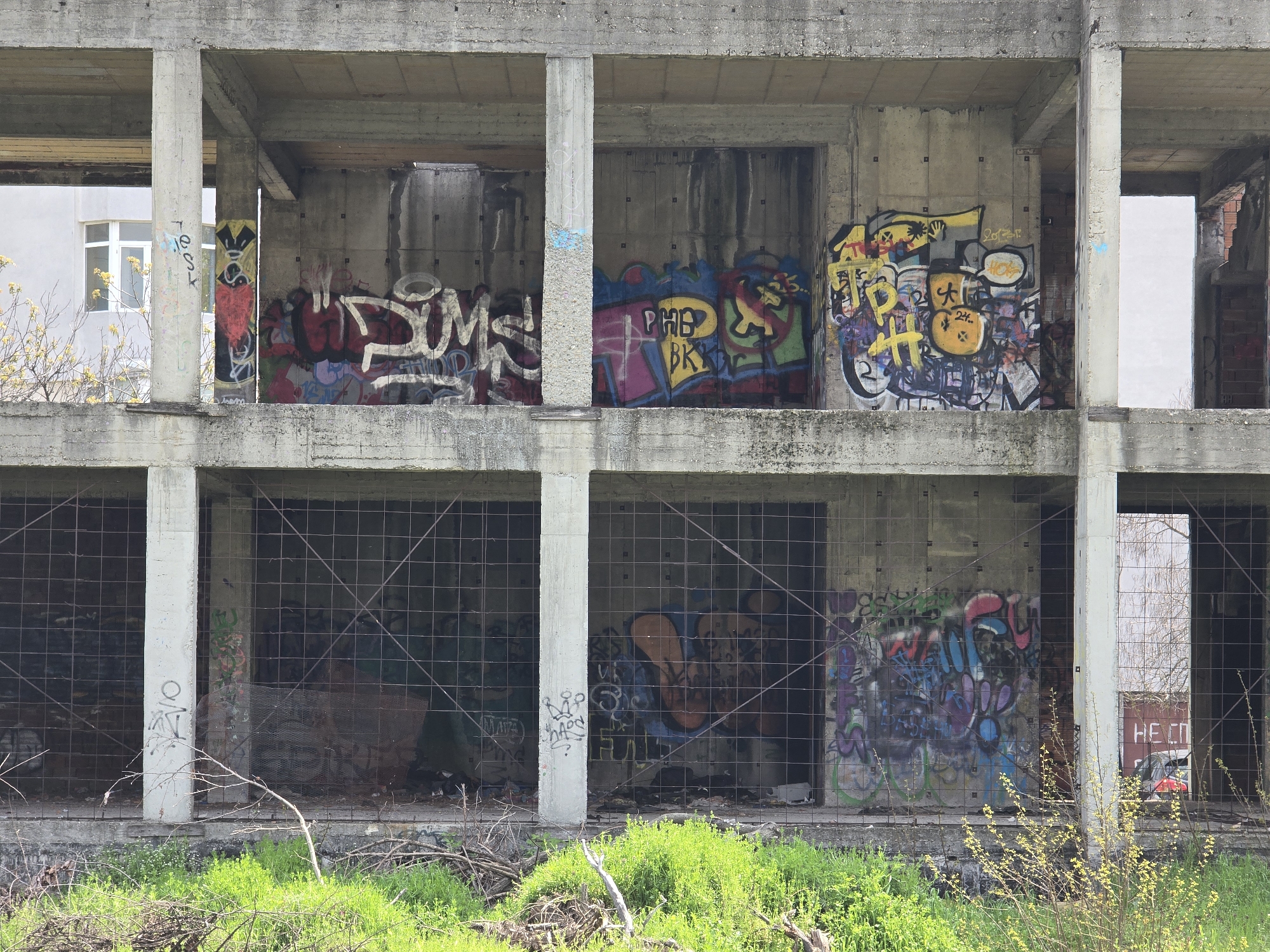 The image shows a concrete building, possibly abandoned, with two stories of open, rectangular bays. Each bay is filled with graffiti on the interior walls. The lower bays are covered with a wire mesh. In the foreground is green grass and some small shrubs. In the background, visible through the bays, are other buildings, trees, and a red car. The location appears to be in a city, but the specific city is unknown.