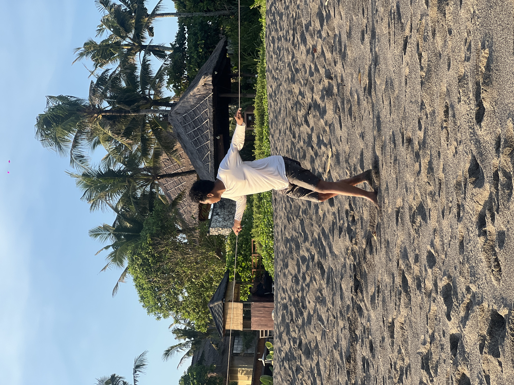 A person stands on a sandy beach in Mengwi, Indonesia. The person is wearing a long-sleeved white shirt and dark shorts, holding a thin rope with one hand extended upwards. The beach is bordered by tropical vegetation, including palm trees and thatched-roof structures. The sky is clear and blue. The sand is dark, and footprints are visible.