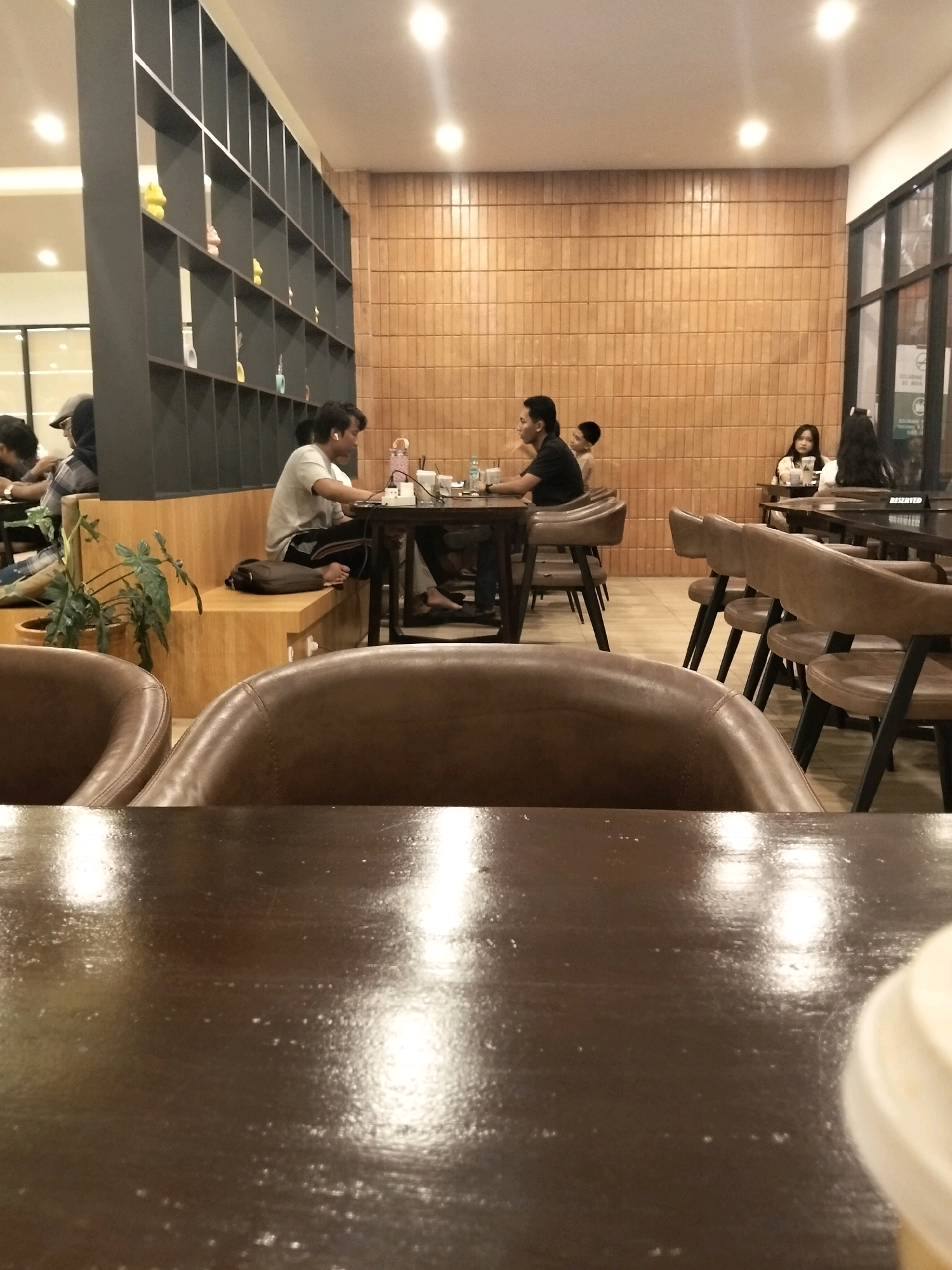 The image shows an interior scene of a cafe, likely located in Arang Limbung, Indonesia. In the foreground, a dark, reflective table surface occupies the bottom portion of the frame. Behind the table, several brown leather chairs are visible. Further back, patrons are seated at tables.
On the left, a person wearing a gray t-shirt and dark pants is seated at a table, appearing to work on a laptop. A potted plant with large, green leaves is situated near the seating area. To the right of this individual, another person is seated at a table with a dark t-shirt and dark pants. A young boy is visible standing next to this person, looking towards the right.
In the background, against a wall with horizontal wooden paneling, several other individuals are seated at tables. On the far right, two women are seated at a table with a "RESERVED" sign visible. The cafe features a dark shelving unit on the left side, filled with various decorative items. The lighting in the cafe appears to be artificial, with ceiling lights casting a warm glow. The overall atmosphere suggests a casual dining or work environment.
