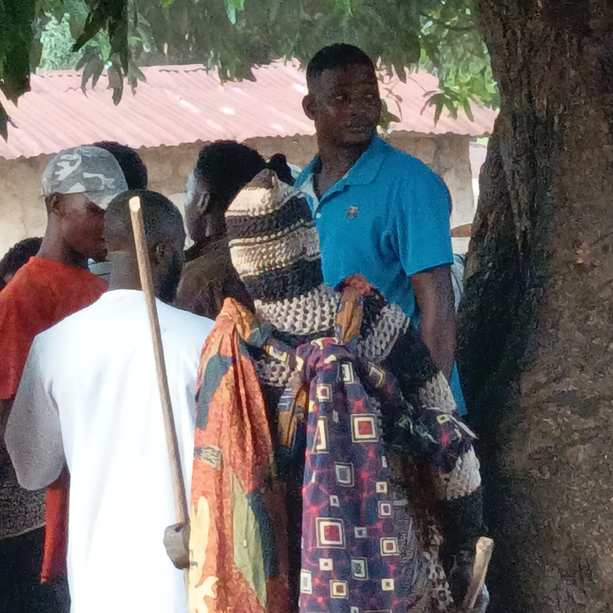 In Akwanga, Nigeria, a group of people gather near a tree. A man in a blue polo shirt stands near the tree, looking away from the camera. Another man is wearing a white shirt and holding a long stick in his left hand. Beside them is an object draped with colorful fabrics. In the background, other individuals are present, including one wearing a camouflage cap and another in a red shirt. A building with a red roof is also visible in the background.