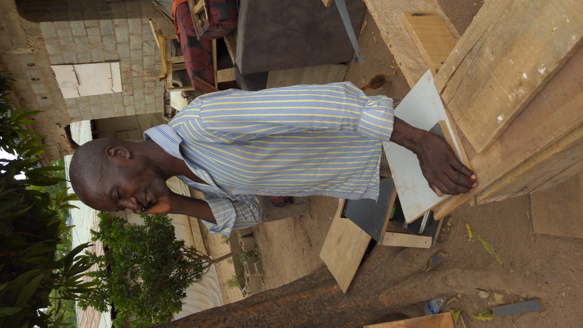 An adult man with dark skin, short hair, and a light beard is captured in an outdoor setting in Masaka, Nigeria. He is wearing a light blue and yellow striped long-sleeved shirt and is actively engaged in a phone call, holding a dark mobile phone to his right ear. He has a broad smile, indicating a pleasant conversation. His left hand rests on a light-colored wooden surface next to stacked wooden planks, suggesting he might be in a woodworking area or near unfinished furniture.
The immediate surroundings feature various wooden materials, including planks and what appear to be parts of unfinished structures or furniture pieces. A hand saw is visible near a dark cushion or sofa in the mid-ground. The ground is unpaved, consisting of dirt and scattered leaves, with a plastic bottle also visible. A small brown chick can be seen wandering on the ground near the wooden items.
In the background, there is an unfinished wall constructed from grey concrete blocks. Red graffiti-like text, "SAL-MAN," is written on one of these blocks. Lush green foliage from trees and bushes frames the left and central-right parts of the background. The scene appears to take place during the daytime, under natural light.