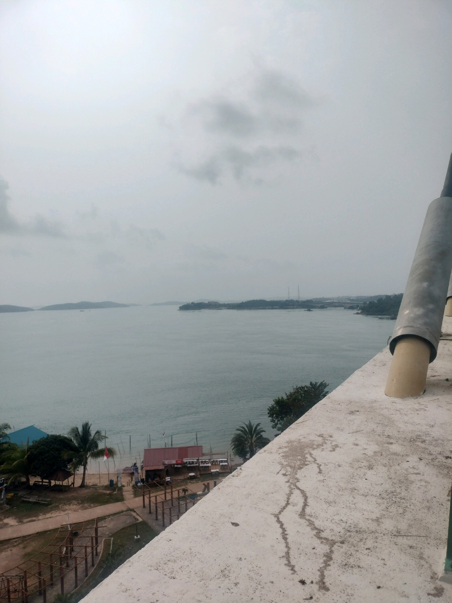 The image captures a panoramic view of a coastline in Batam, Indonesia, on an overcast day. In the foreground, a textured concrete structure, possibly part of a building or observation deck, occupies the lower right portion of the frame, with a large cylindrical pipe extending upwards. The midground reveals a tranquil sea with several islands dotting the horizon. Along the shoreline, a small settlement is visible, featuring a red-roofed building with tables and chairs outside, suggesting a restaurant or cafe. Several palm trees and other greenery are scattered across the sandy beach, with a few small boats moored in the water. The background is dominated by a vast expanse of sea and the hazy outlines of distant islands and structures, possibly including bridges. The sky is a uniform grey, with diffused light indicating a cloudy or foggy atmosphere. No people are clearly visible in the scene.