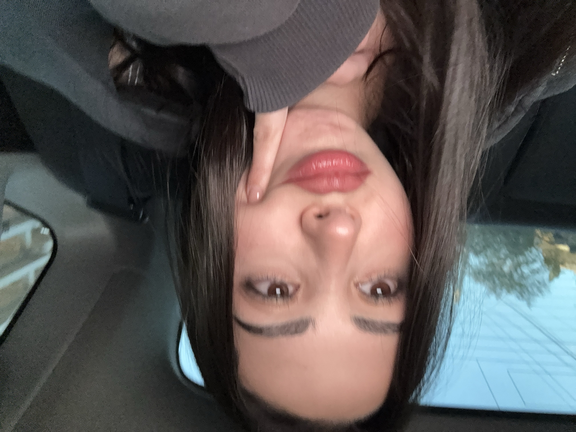 A young woman is taking a selfie indoors, possibly in a car. The camera angle is upside down, revealing her face, dark hair, a grey sweatshirt, and a hand touching her face. Outside the car window, there are trees and a light blue sky, suggesting a daytime setting. There are no specific clues to place the location in Yerevan, Armenia.