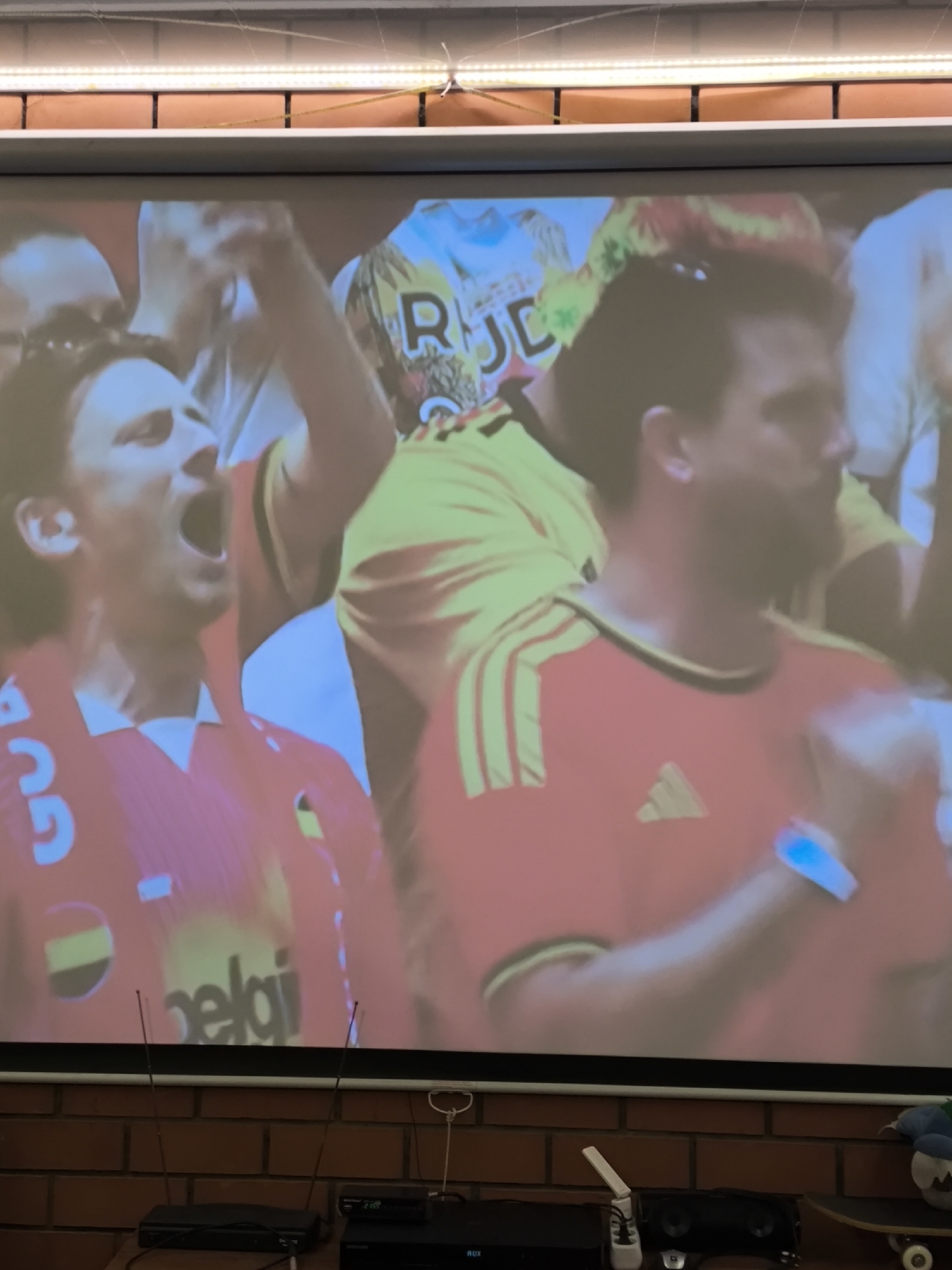 In Pokrov, Russia, a large projection screen displays an image of three men in red and yellow jerseys, seemingly at a sporting event. The man on the left, with dark hair, is shouting with his mouth open wide, wearing a red jersey with a white collar and blue lettering on the front. To his right, another man, also in a red jersey with yellow stripes on the sleeves and an Adidas logo, looks towards the right. A third man, partially visible behind them, wears a yellow jersey with black accents and a white logo. The projection is set against a backdrop of brick walls. Below the screen, electronic devices, including a black box with antennas and another device with a digital display, are placed on a brick surface.