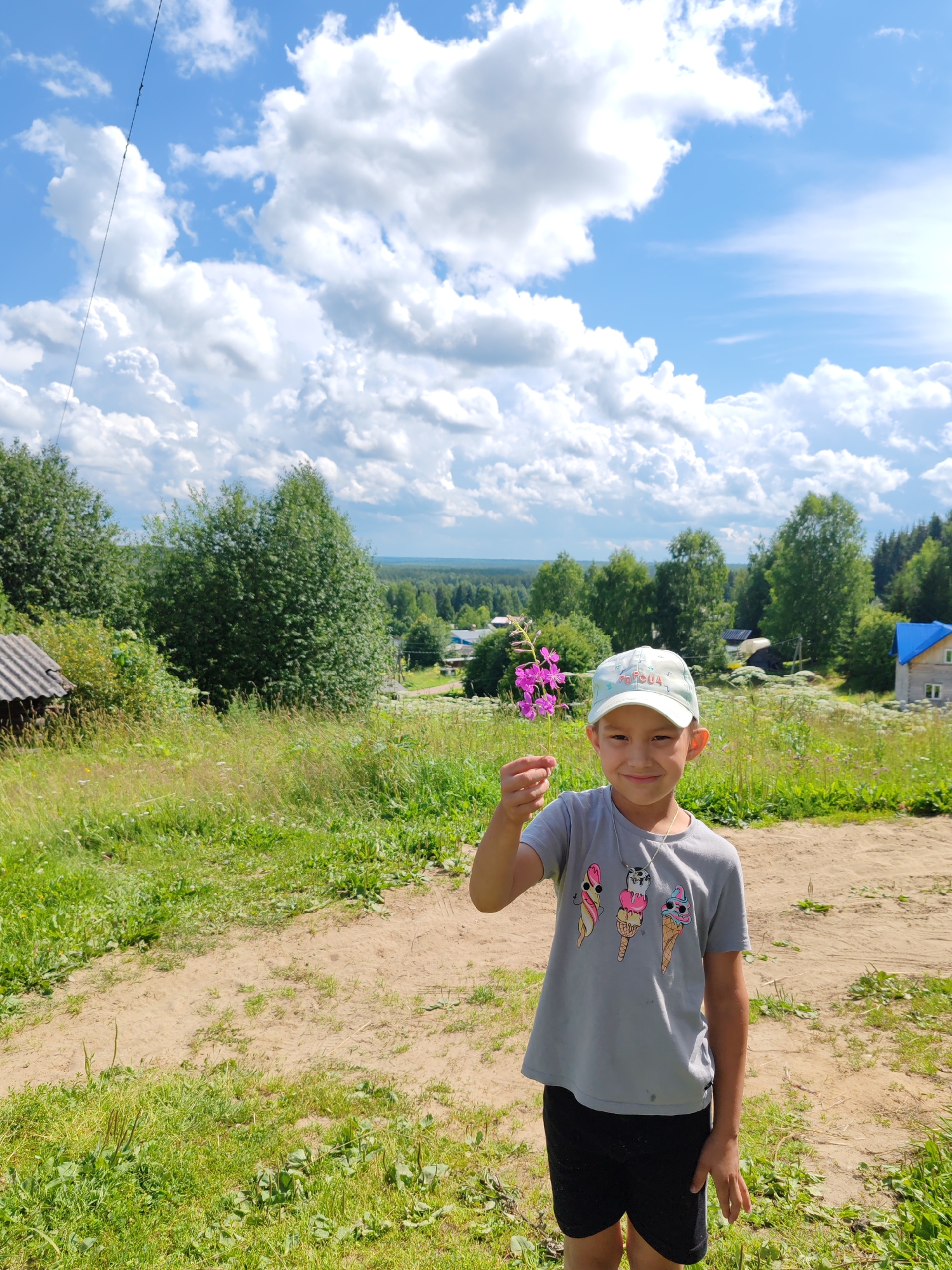 A young boy with short dark hair and a light blue baseball cap stands in the foreground, holding a small branch with purple flowers. He is wearing a grey t-shirt with colorful ice cream cone graphics and black shorts. The boy is smiling and looking at the camera. Behind him, a dirt path leads away from the viewer, bordered by green grass and tall wildflowers. In the background, a wooded landscape stretches out under a partly cloudy blue sky. A small, rustic wooden structure with a corrugated metal roof is visible to the left of the path, and a blue-roofed house is situated further back in the middle ground. The scene appears to be in a rural area, possibly the village of Chukhlem, Russia, given the architectural style and natural environment.