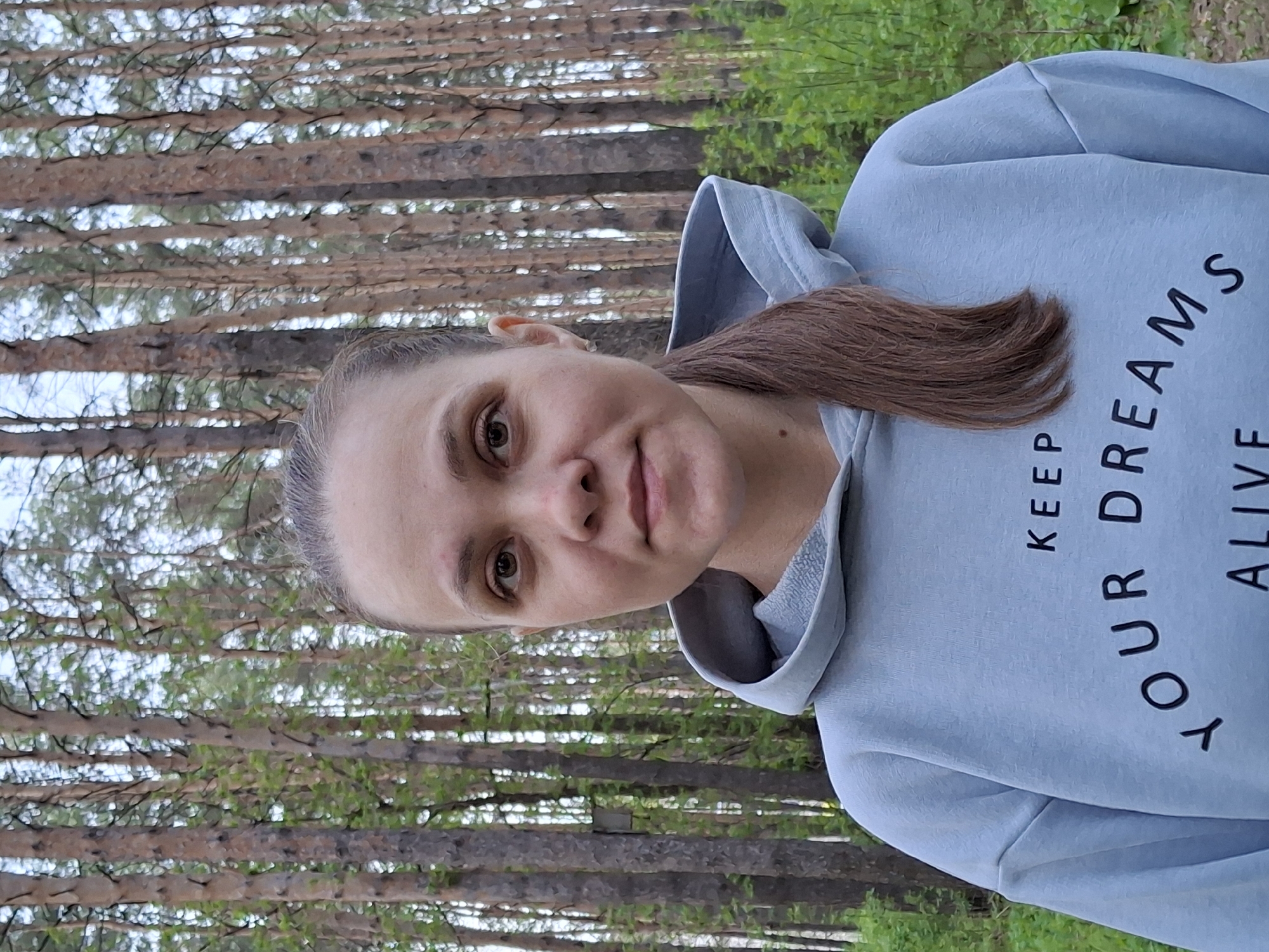 A woman stands in a forest, likely near Chaikovsky, Russia, given the prevalence of pine trees. She is wearing a light blue hoodie with the phrase "KEEP YOUR DREAMS ALIVE" printed on it. Her brown hair is pulled back, and she has light skin. The background consists of closely spaced tall pine trees with green foliage near the ground.