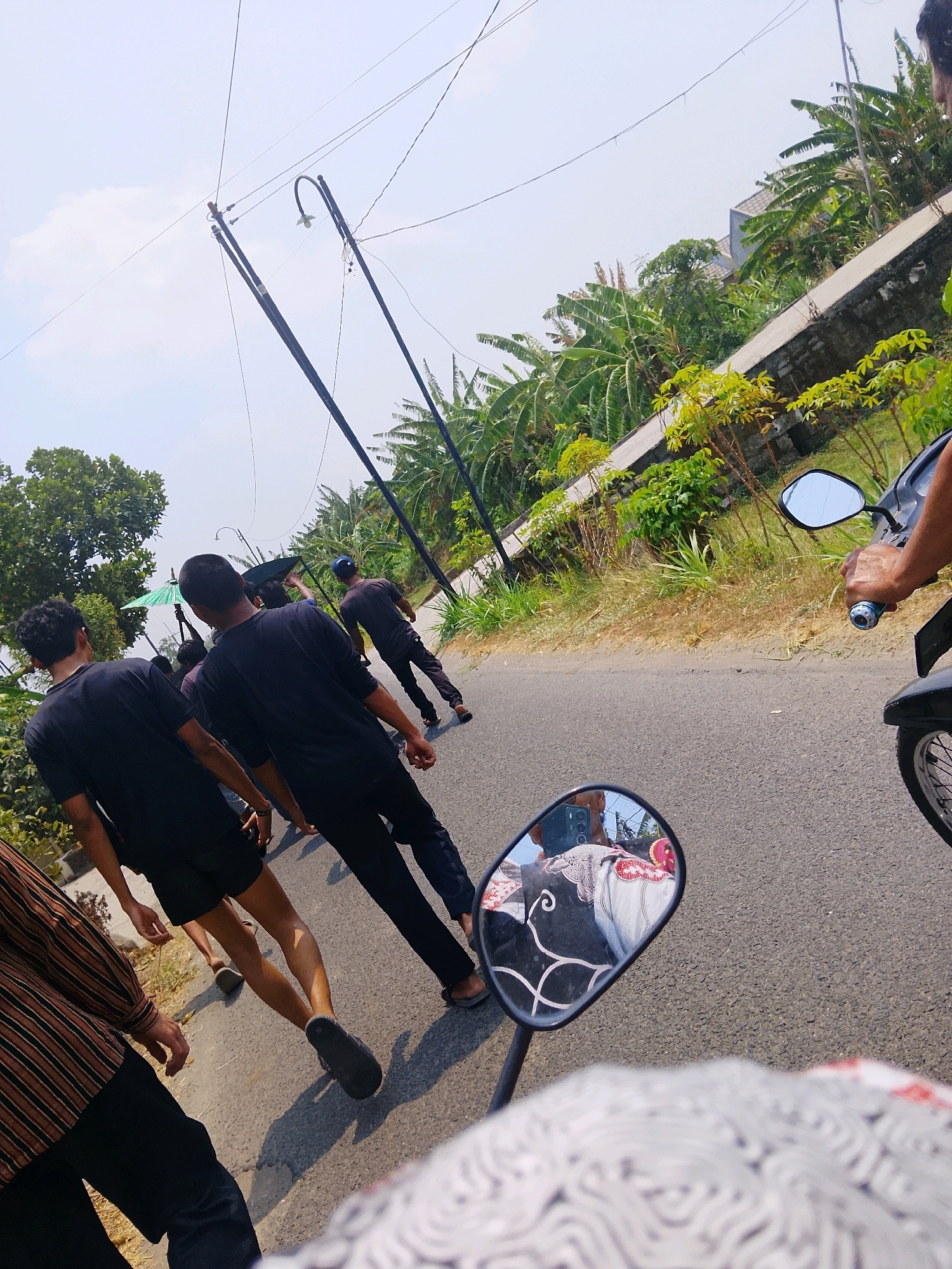The image shows a group of people walking on a road in Kepuh, Indonesia. The people are wearing casual clothes, and some of them are carrying umbrellas. The road is surrounded by trees and vegetation, and there are some buildings in the background. The sky is clear and sunny. The image is taken from a high angle, and it is difficult to see the faces of the people. The overall scene appears to be a peaceful gathering in a rural area.
