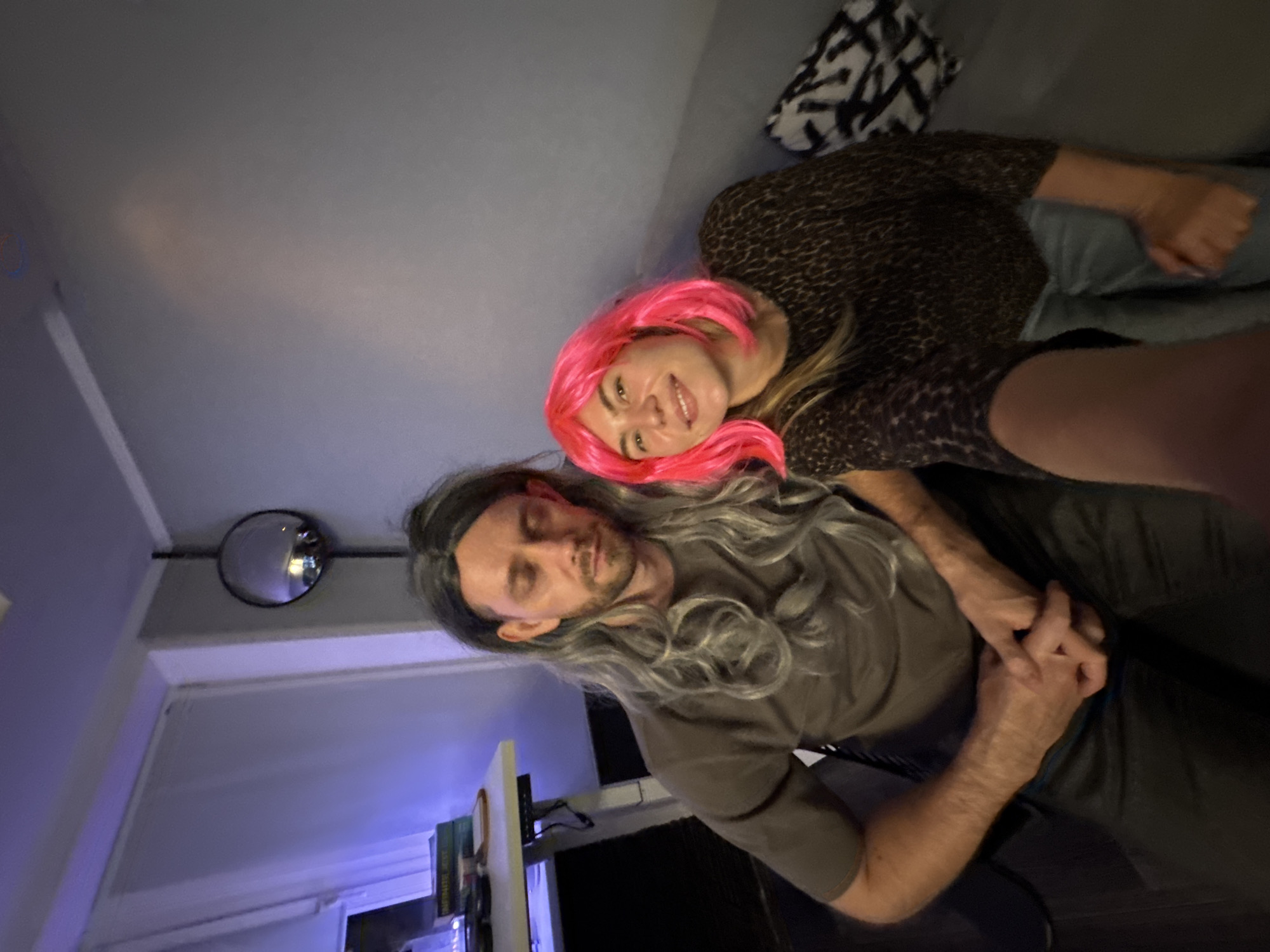 Two individuals, one with long grey hair and a grey t-shirt, and another with bright pink hair and a leopard-print top, are posing for a selfie. The person with grey hair is sitting and appears to be wearing dark pants. The person with pink hair is sitting next to them, wearing light blue jeans. They are indoors, in what appears to be a living space. In the background, there is a grey wall, a desk with a laptop, and a round mirror hanging on the wall. The lighting in the room is dim, with a purple hue coming from the left side of the image.