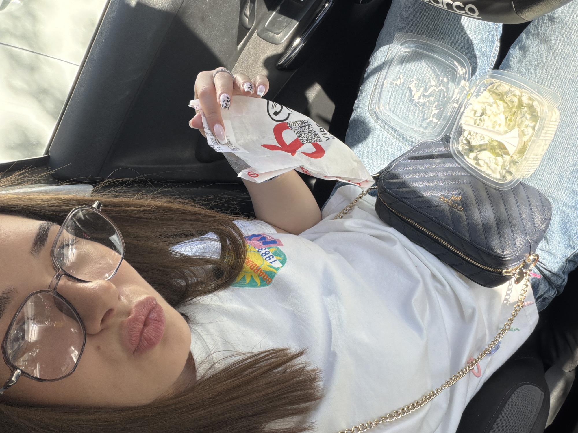 The image shows a person in a car holding a bag of food from KFC. The person is wearing glasses and a white shirt with a colorful logo that reads "Guardian 1986". They have manicured nails with black and white designs. On their lap is a small black purse with a gold chain strap, an opened container of coleslaw, and the lid to the container. The person is wearing light blue jeans. It is daytime and the car appears to be parked. The location is most likely not in Sofia, Bulgaria as the image features KFC, a brand not present in Bulgaria.