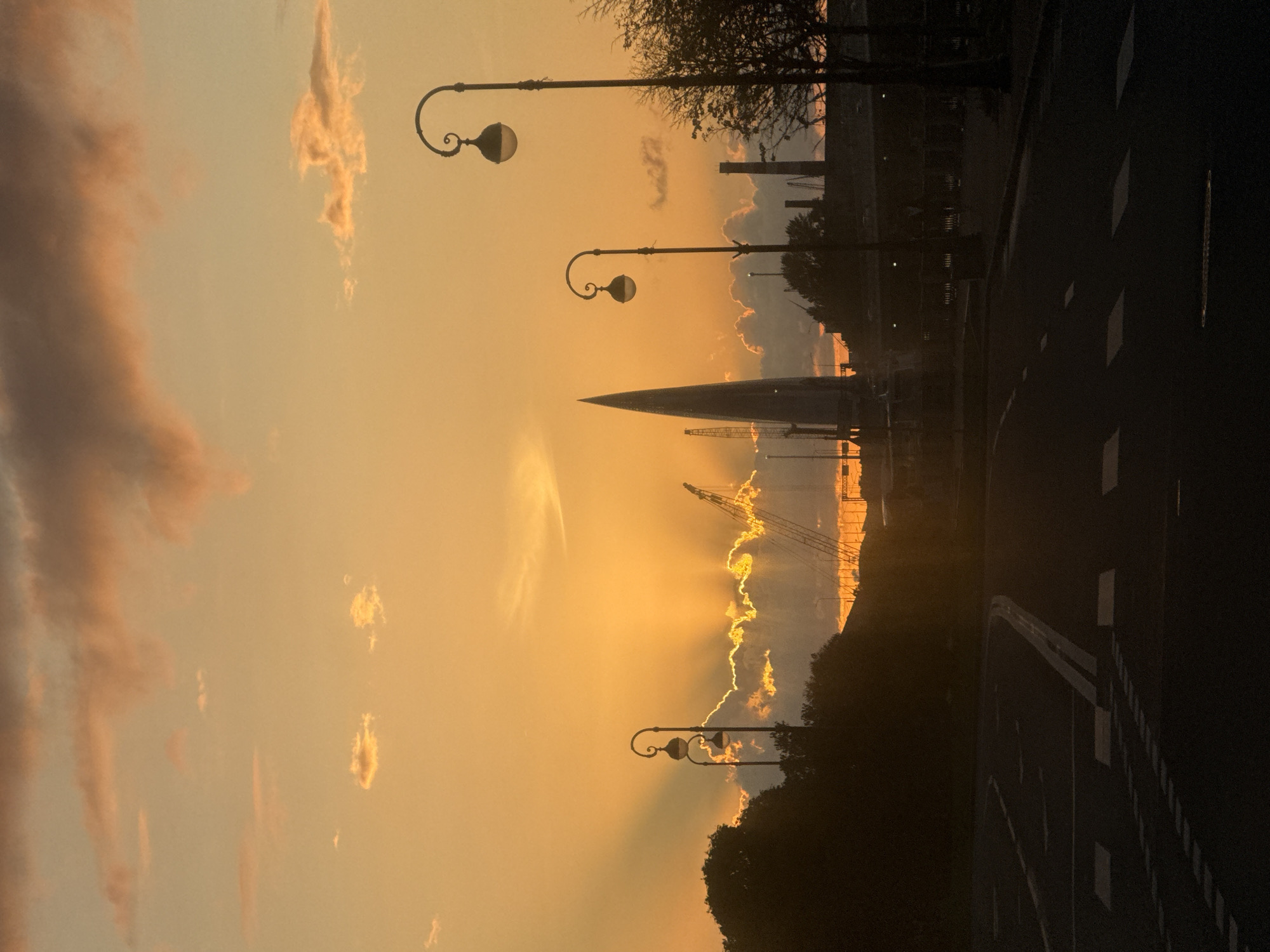 The image depicts a sunset over the city of Saint Petersburg, Russia, likely from a vantage point overlooking the Neva River or one of its tributaries, given the presence of industrial structures and a prominent spire. The sky is a vibrant orange and yellow, with clouds tinged with pink and orange from the setting sun. Rays of sunlight are fanning out from behind a band of clouds. In the foreground, a dark, silhouetted road with lane markings is visible. Two ornate streetlights stand on the right side of the road, and a third, slightly further away, is on the left. In the middle ground, silhouetted trees and industrial buildings, including chimneys and cranes, are present. A distinctive, tall, pointed structure, resembling a skyscraper or a monument, is visible to the left of the center. The scene conveys a sense of twilight and industrial urban landscape. There are no people or animals visible in the image. The time of day is sunset.