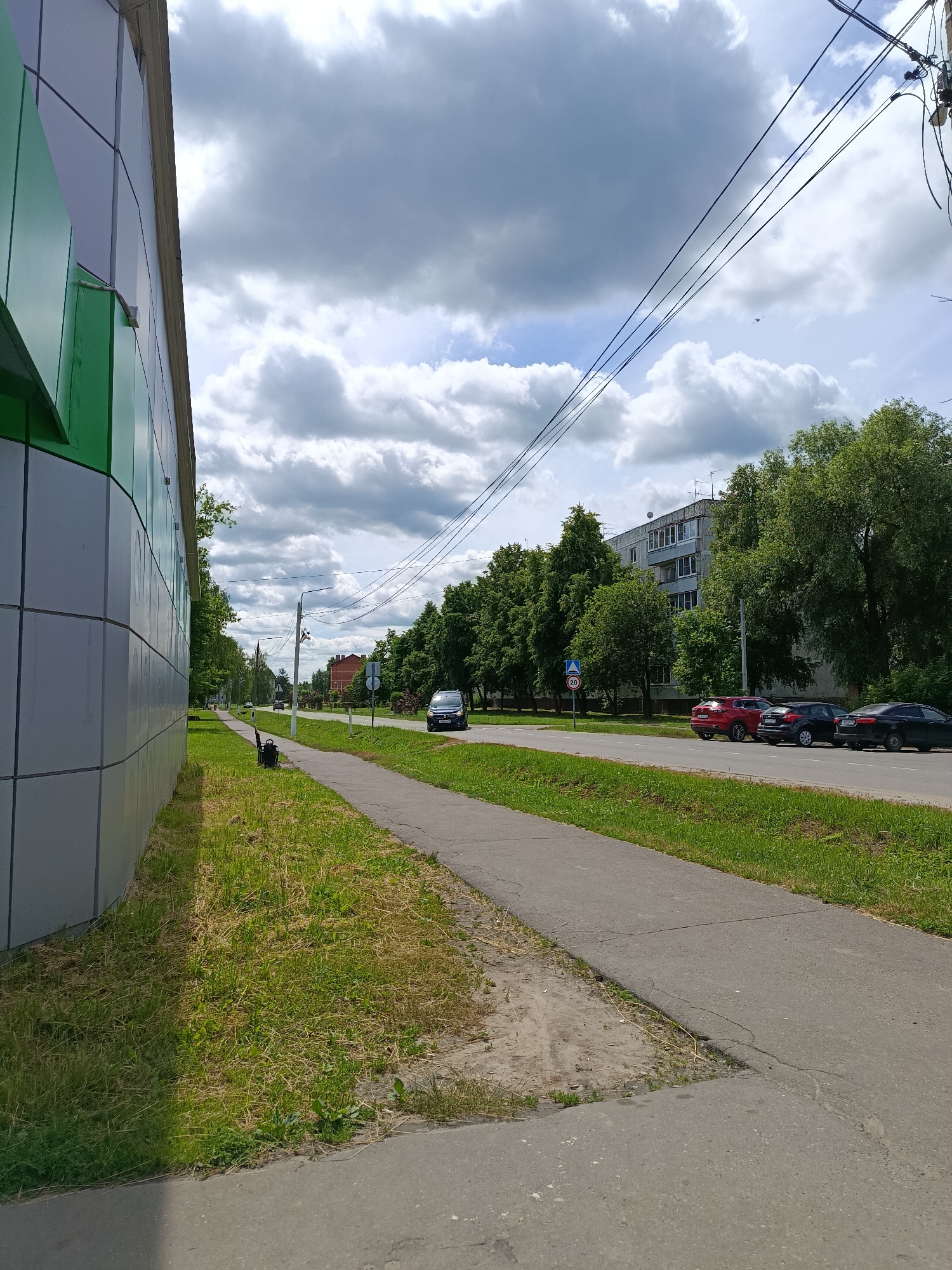 This is a street-level, eye-level, full-shot photograph taken outdoors on a cloudy day. The composition is dominated by a sidewalk and grassy verge on the left, bordered by a modern building with a grey and green facade. The sidewalk extends into the distance, meeting a paved road where several cars are parked and in motion. On the right side of the road, there is a dense line of green trees, behind which a multi-story apartment building is visible. Further down the road, more trees and buildings are present, suggesting an urban or suburban environment. Power lines stretch across the sky, supported by utility poles. A street sign with a speed limit of 20 is visible near the road. The overall scene depicts a typical streetscape with elements of both modern architecture and natural greenery.